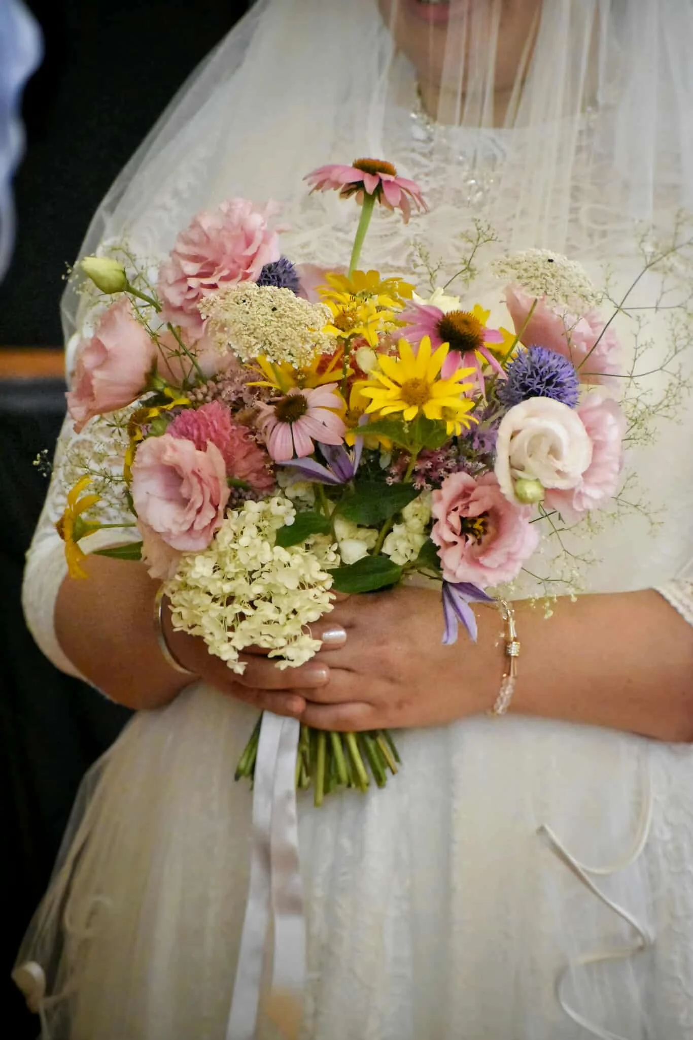 A bride holding a bouquet of colorful flowers, including pink, yellow, purple, and white blossoms, in a wedding dress with lace and a veil.