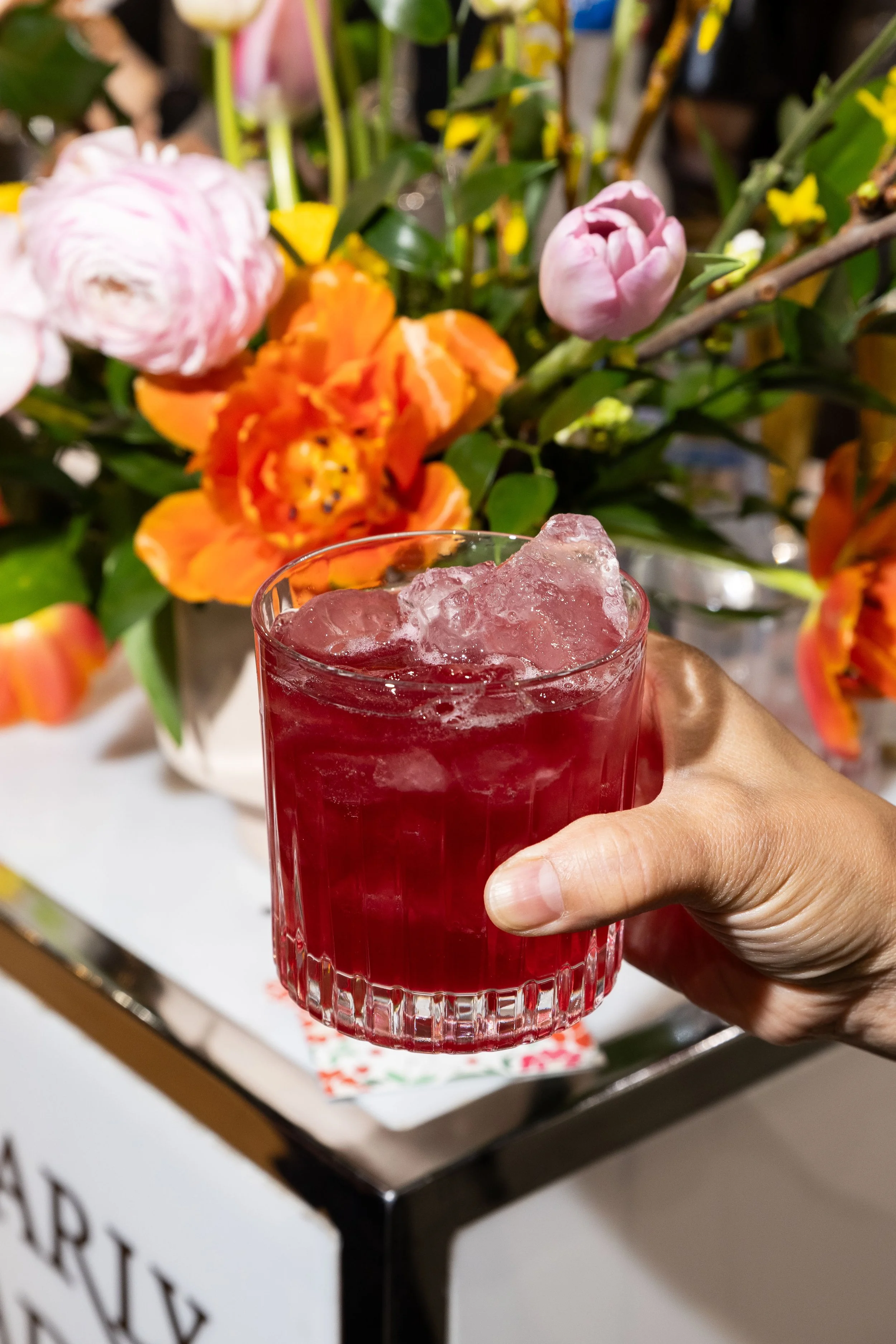 A hand holding a glass of red beverage with ice, in front of a flower arrangement with pink, orange, and yellow flowers.