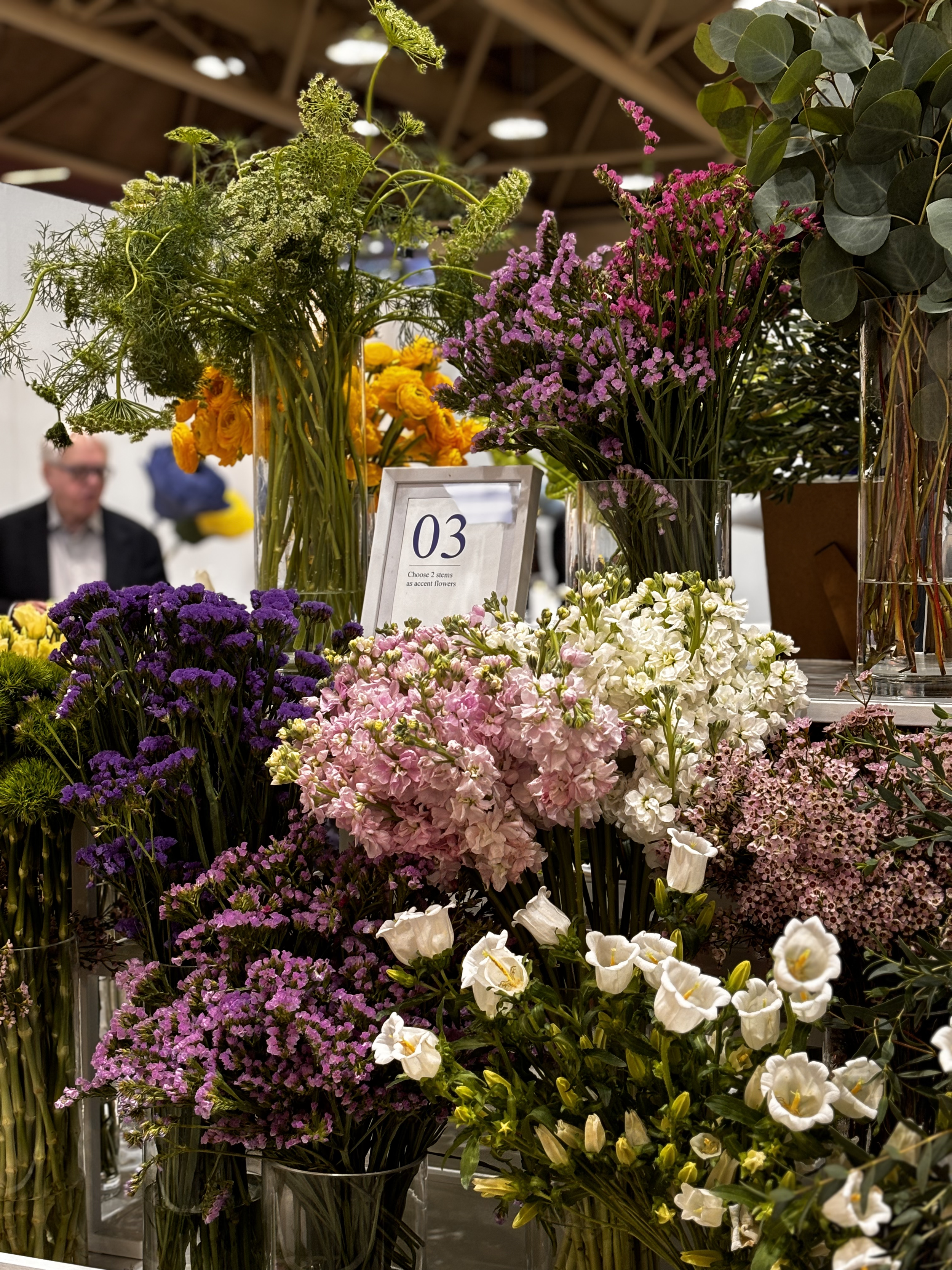 A display of various colorful flowers in vases at a market or flower shop, with a man in the background browsing.