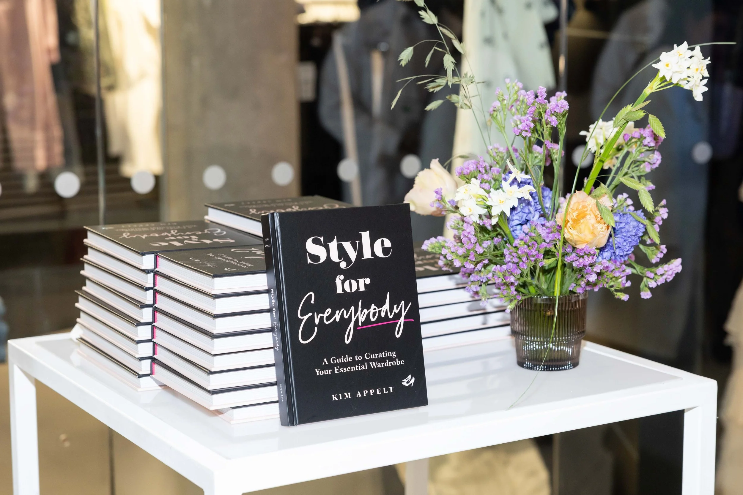 A white table displays a stack of copies of the book "Style for Everybody" by Kim Appelt, with a single copy standing upright in front of the stack. To the right, there is a glass vase filled with purple, blue, and white flowers.