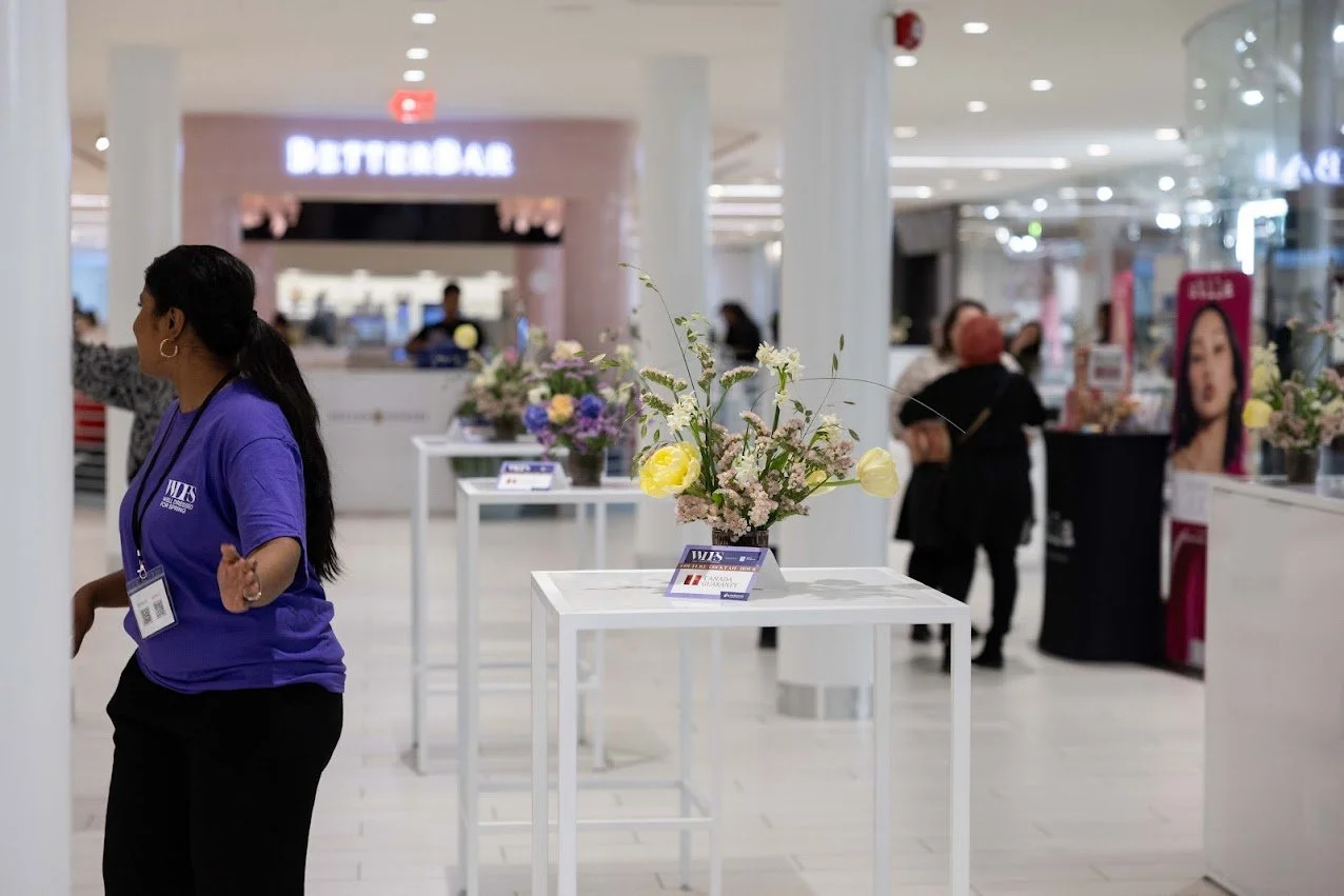 A mall interior with flower arrangements on white tables, and people shopping or browsing. A woman in a purple shirt with a name badge is in the foreground.