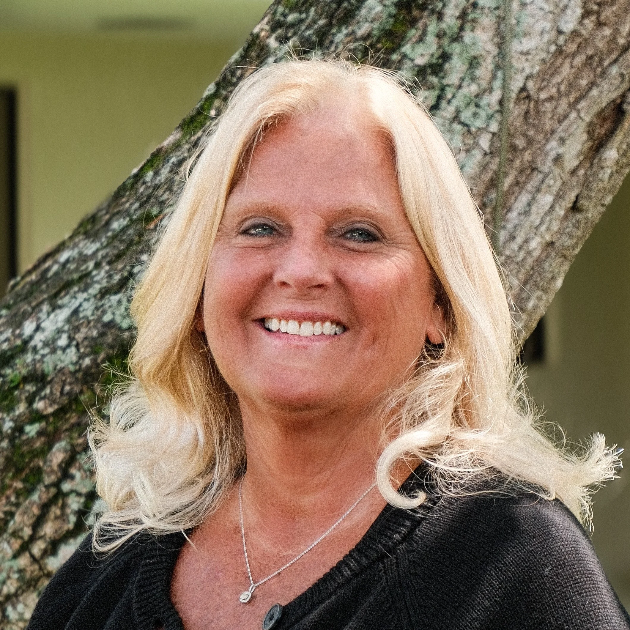 A woman with shoulder-length black hair smiling in a green indoor garden or greenhouse, wearing a blue top.