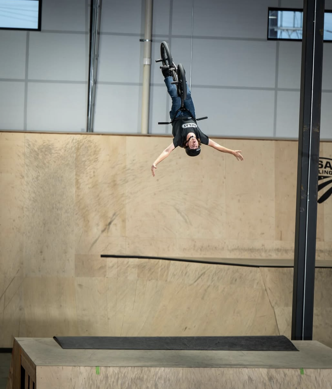 A person in a black t-shirt and helmet performing an acrobatic stunt on a BMX bike inside a skate park with a wooden ramp and a vertical wall.