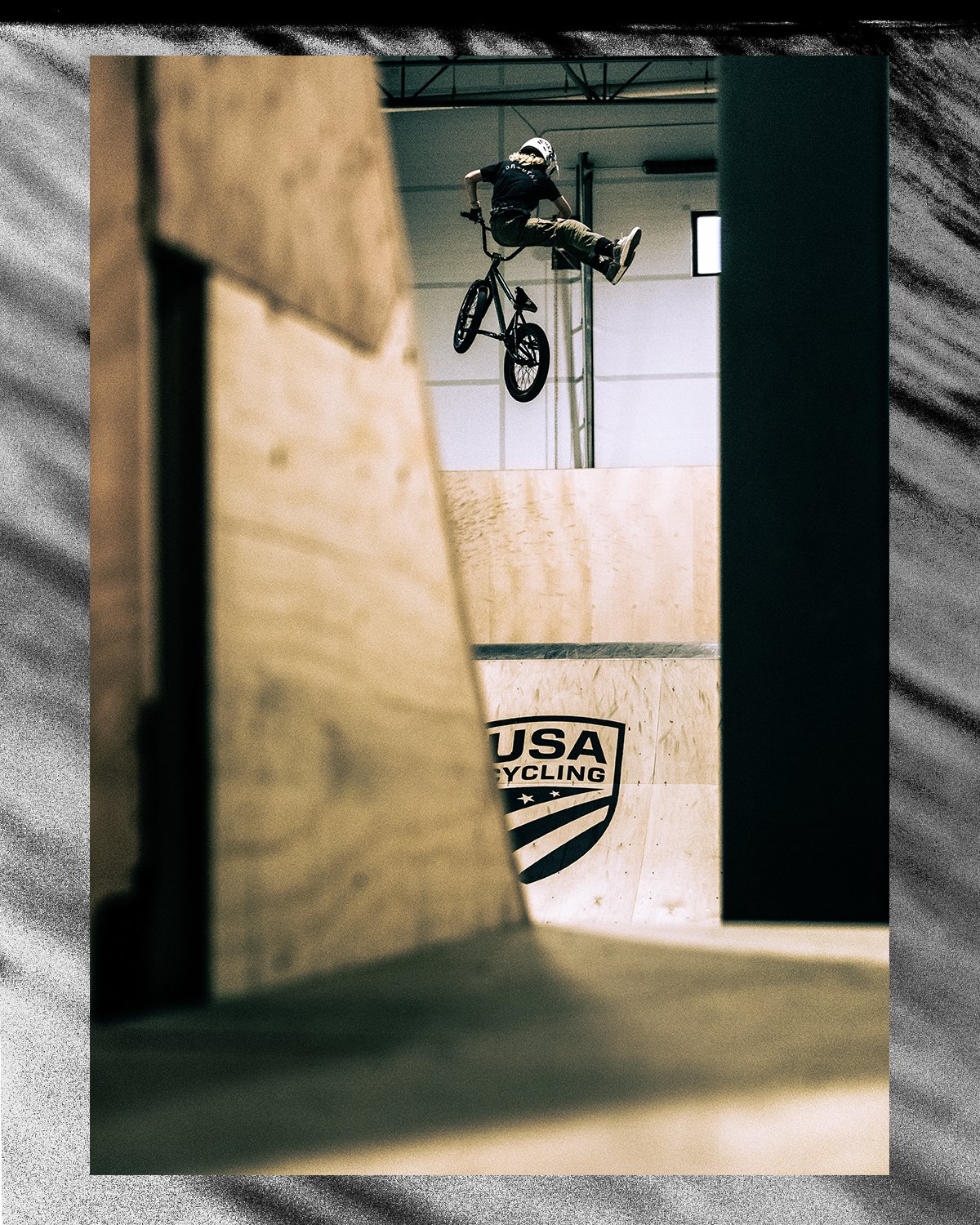 A person performing a BMX trick in an indoor skatepark, mid-air with the bike upside down against a Wood USA Cycling sign.