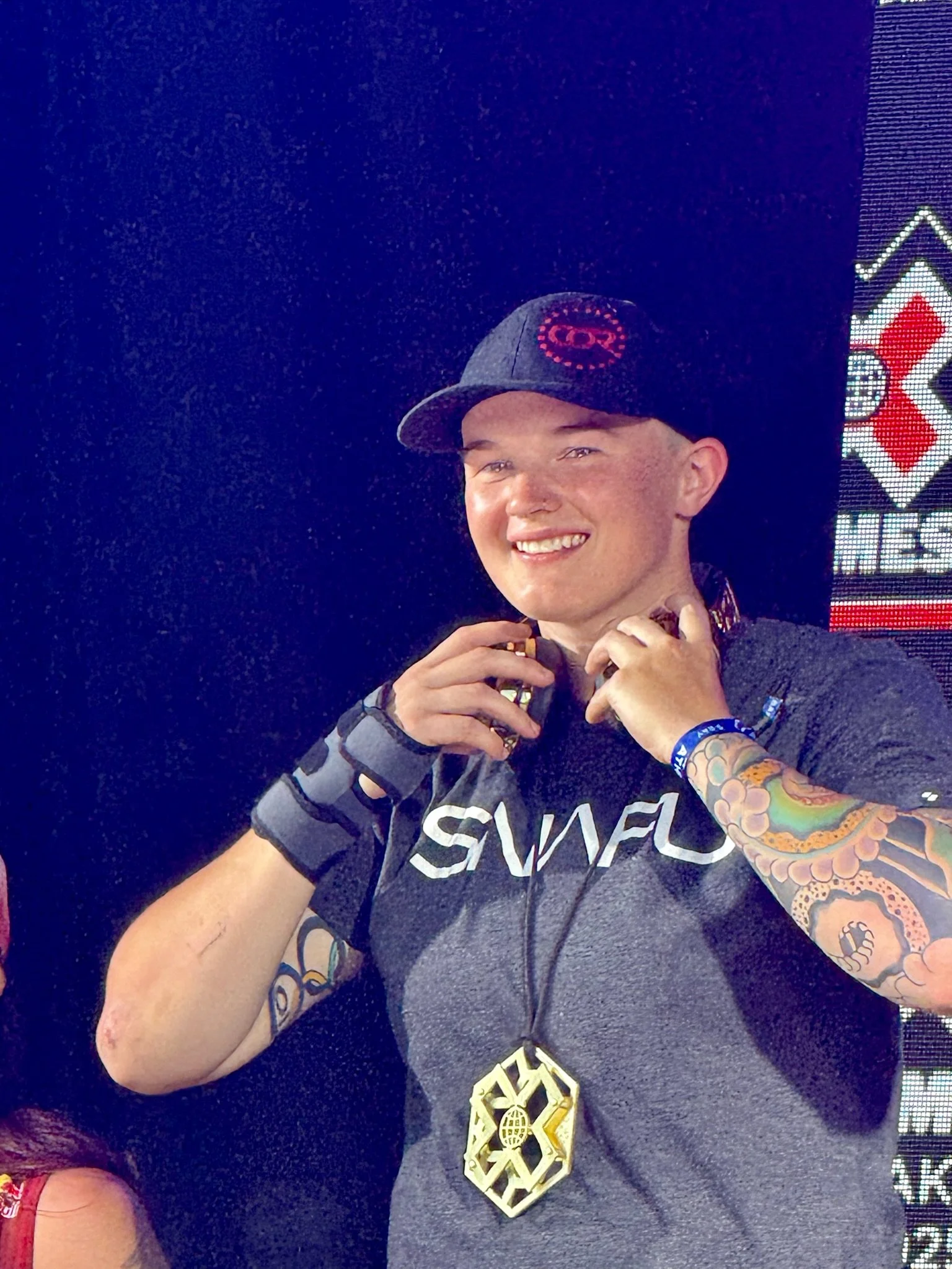 A young man with tattoos on his arm, wearing a black cap, athletic gear, and a medal around his neck, smiling at the camera.