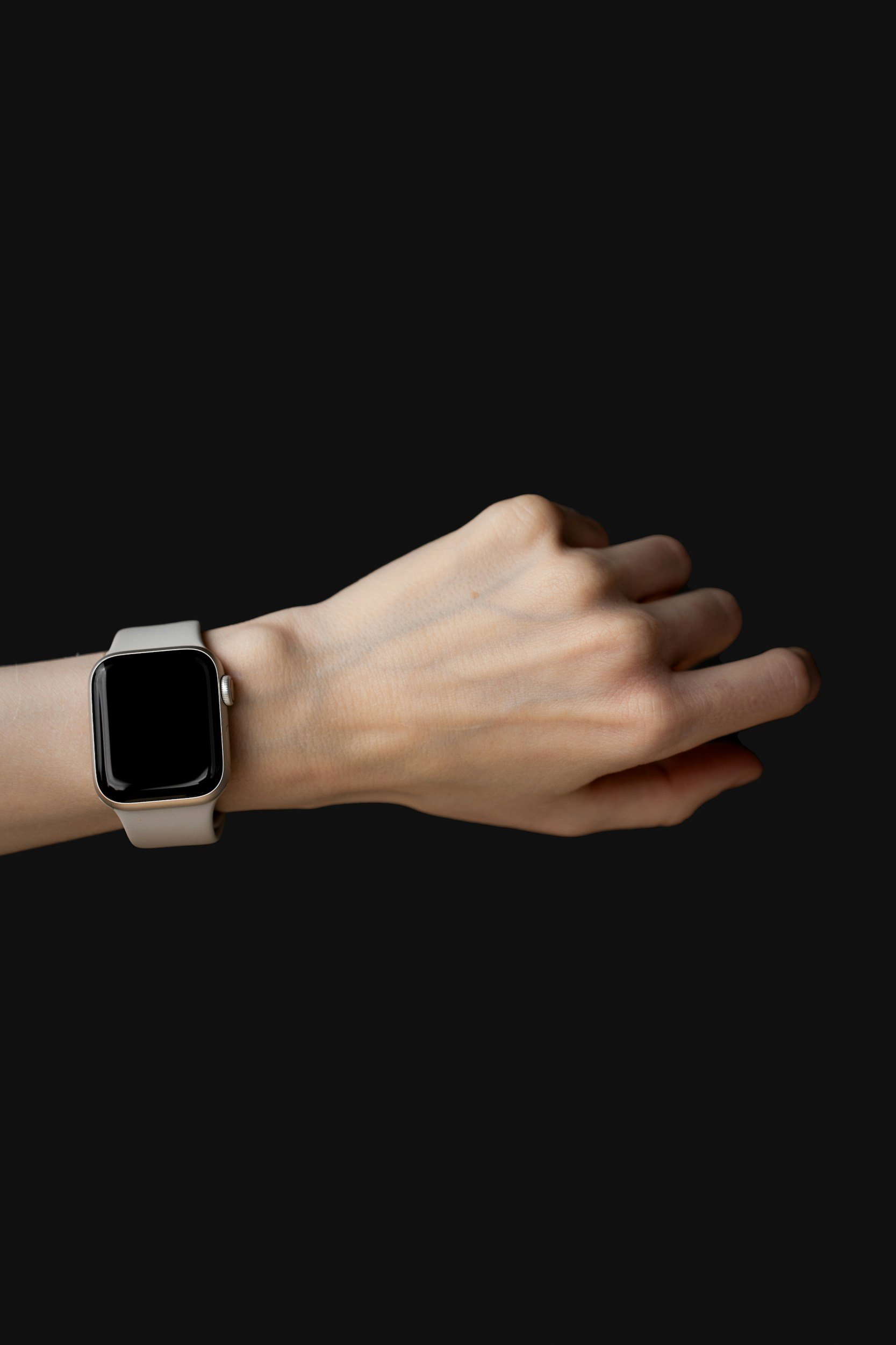 A person's left hand and wrist wearing a smartwatch with a black screen and a light-colored band, against a black background.