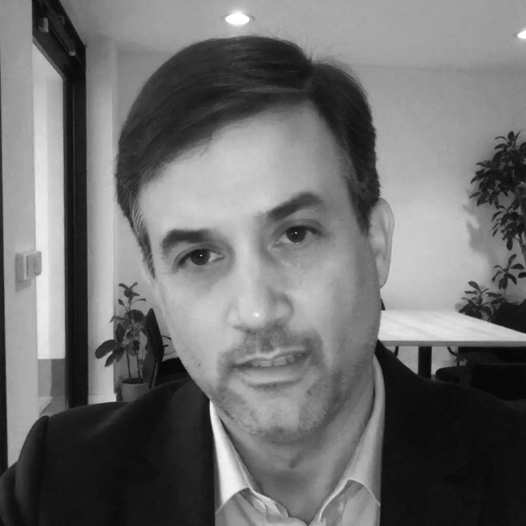 Black and white photo of a man with dark hair, wearing a suit jacket and dress shirt, sitting indoors with plants in the background.