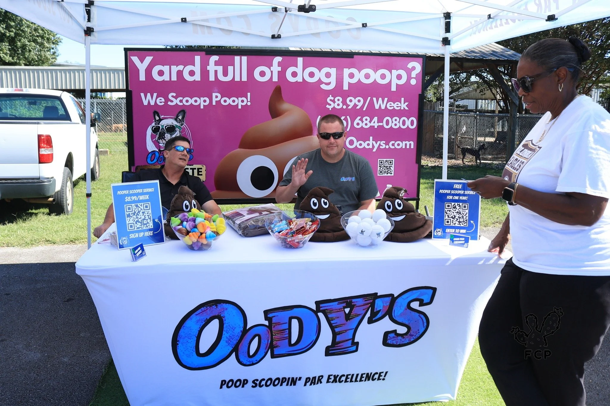 A booth with a banner reading 'Yard full of dog poop? We Scoop Poop! $8.99/Week' is set up outdoors under a canopy, with two men sitting behind a table filled with poop-themed items, including plush poop toys. A woman is standing in front of the booth, looking at her phone. The booth promotes a pet waste removal service called Oodys.