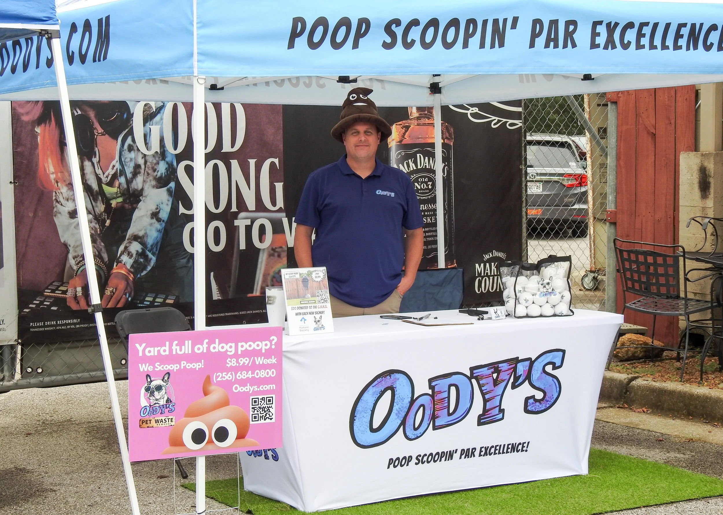 Man standing at an outdoor booth with a blue canopy tent promoting pet waste scooping services. The booth has a white tablecloth with  logo and slogan, and there is a pink sign advertising dog poop scooping for $8.99 per week. The man is wearing a blue polo shirt and a poop emoji hat, smiling at the camera.