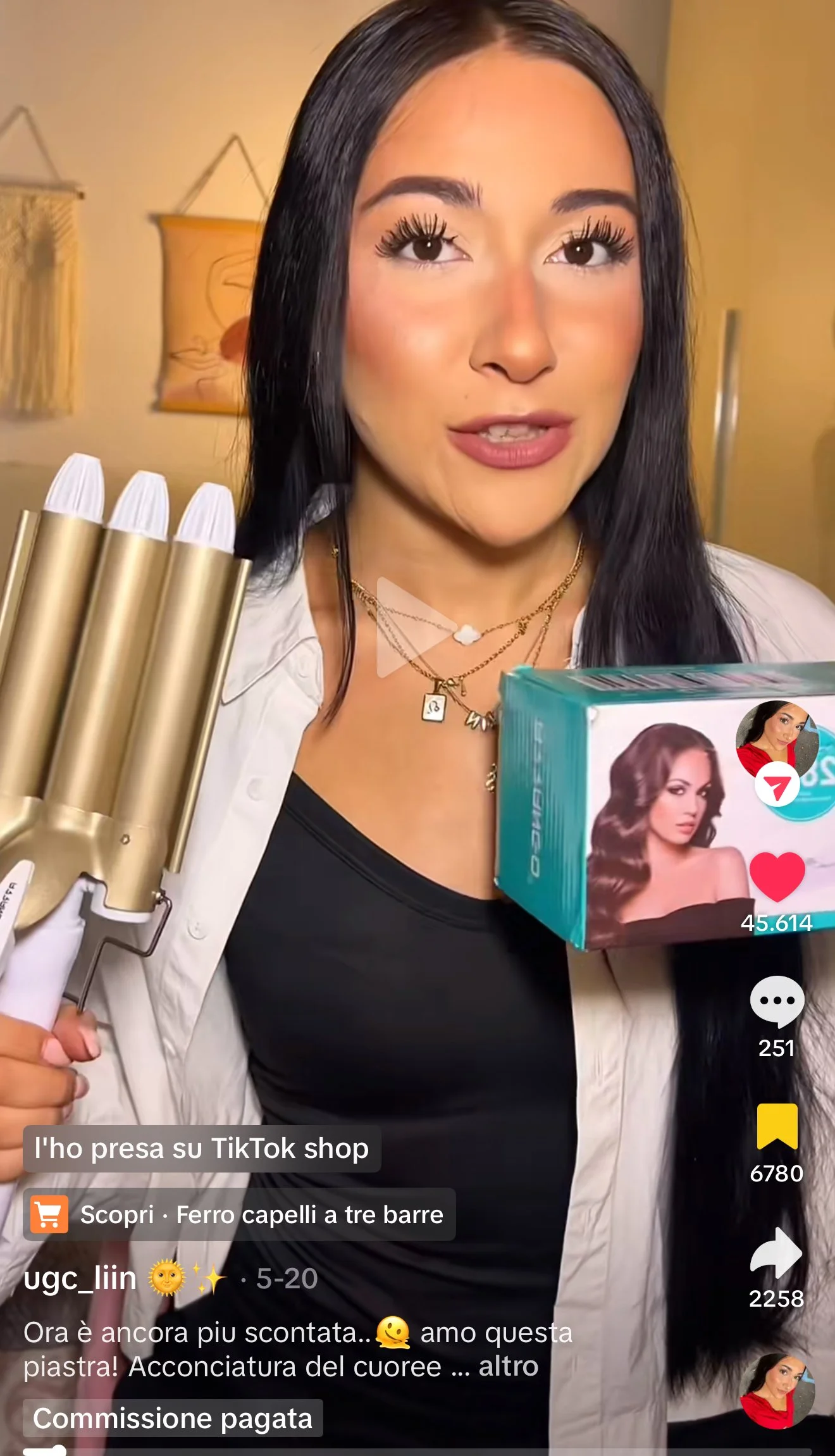 Donna con capelli neri lunghi, indossa una maglietta nera e una giacca chiara, tiene in mano una piastra per capelli a tre barre e mostra una scatola di prodotti per capelli.