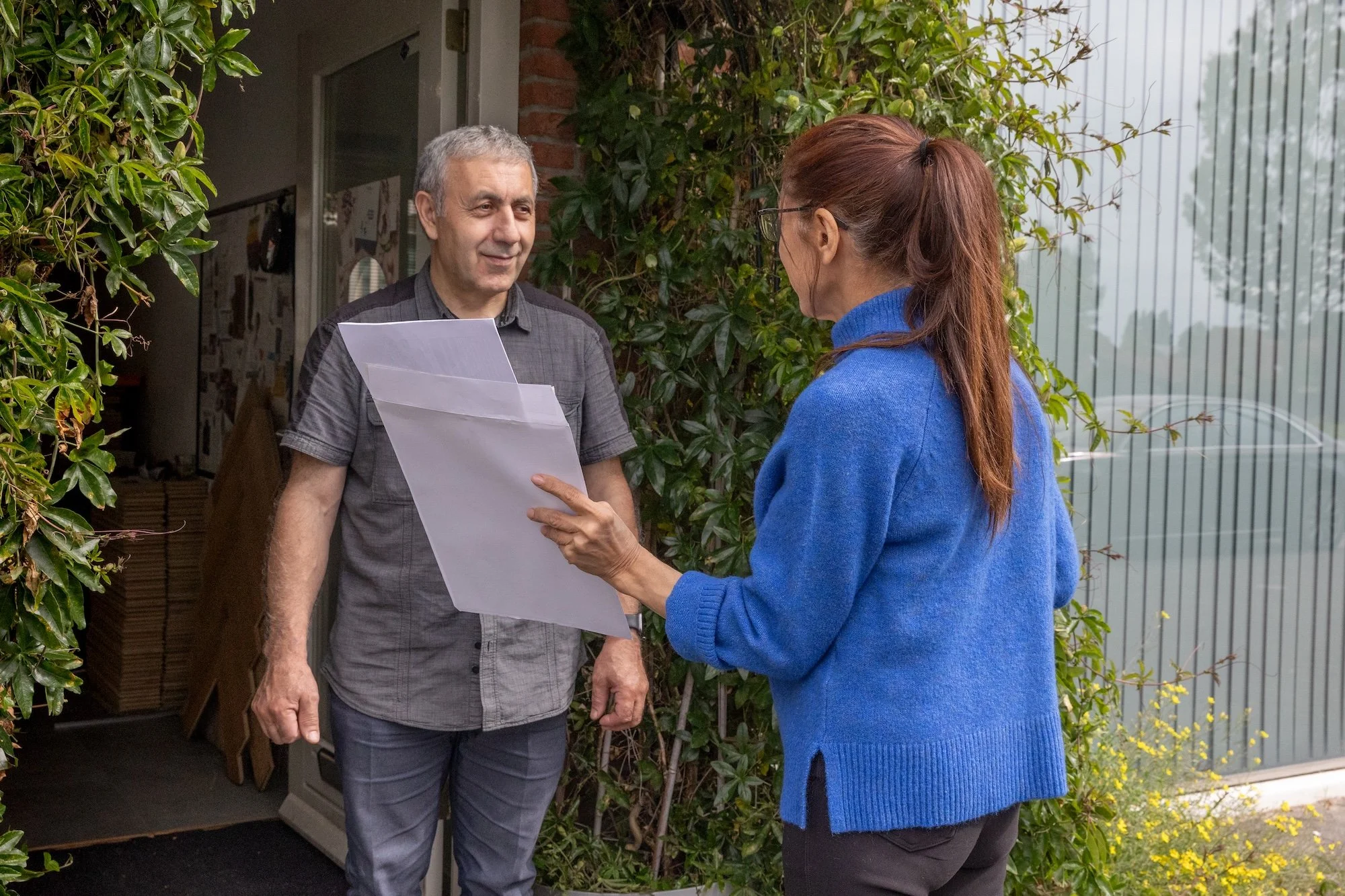 Man met een vrouw voor woning, vrouw houdt papieren in haar hand, de deur staat open.