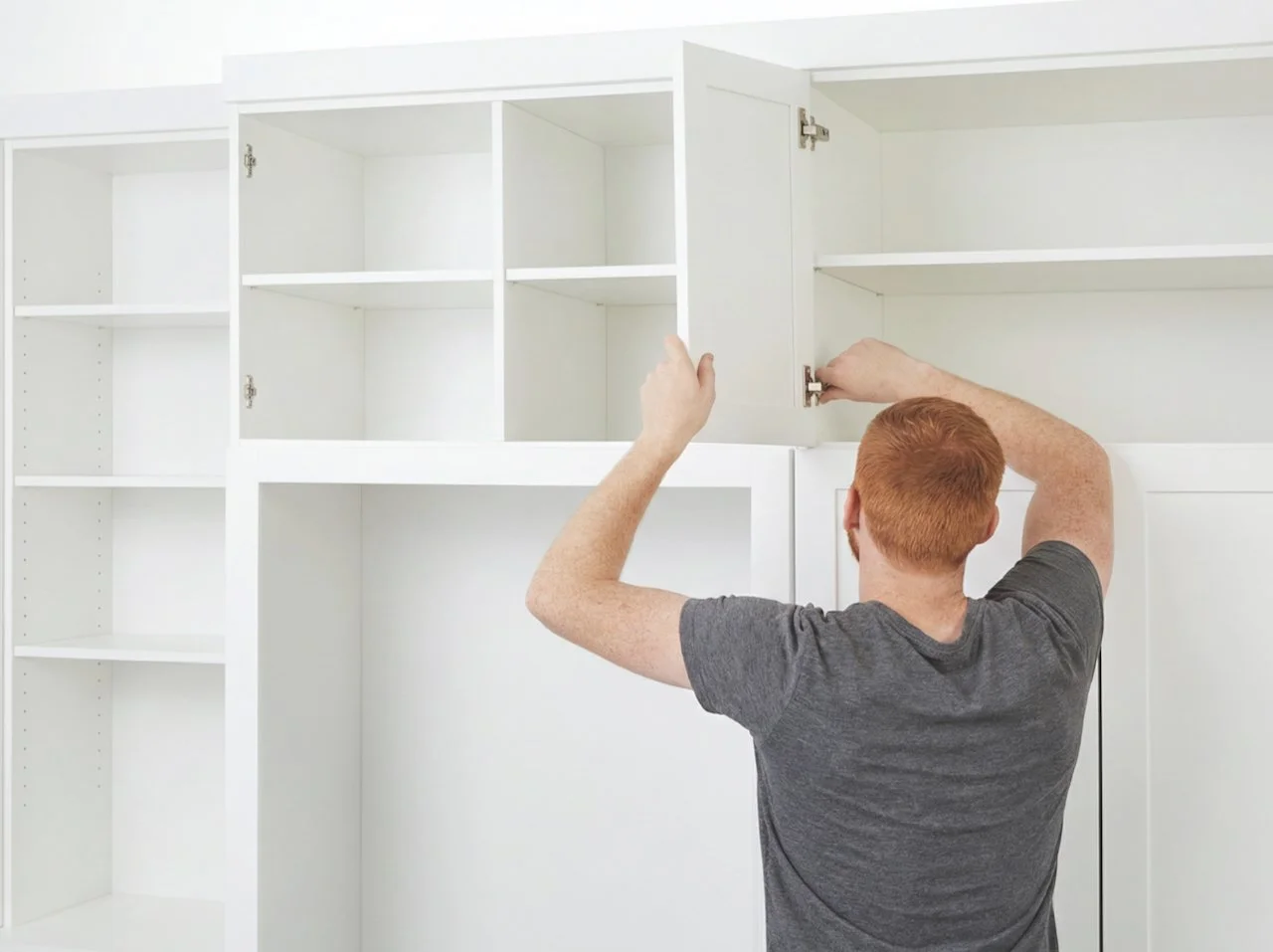 A man with red hair is installing or repairing cabinet doors inside a white built-in cabinet with multiple sections.