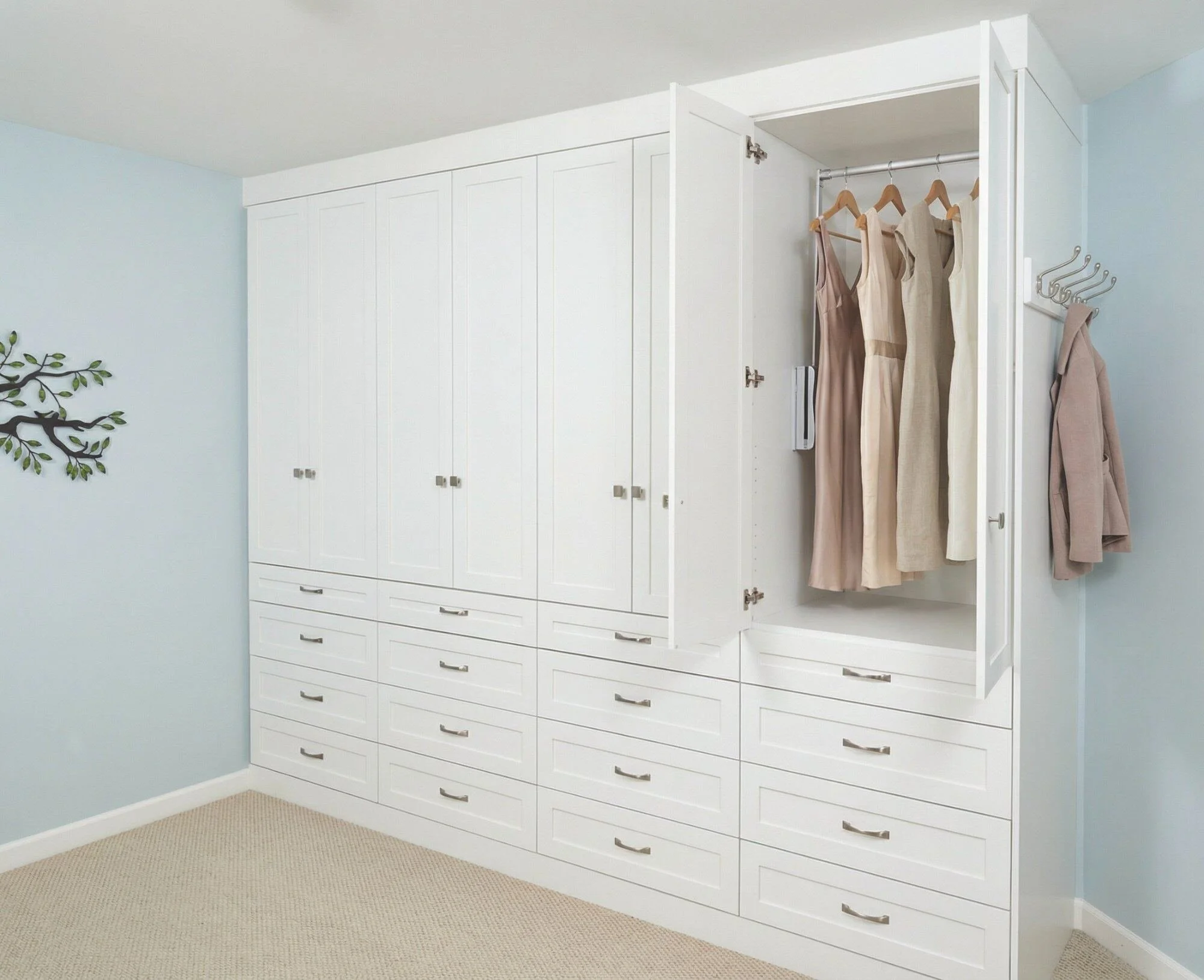 White built-in wardrobe with open doors revealing hanging clothes and drawers, in a light blue room with beige carpet and a decorative wall art