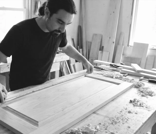 A man in a black shirt inspecting a wooden door in a woodworking shop surrounded by woodworking tools and wood pieces.