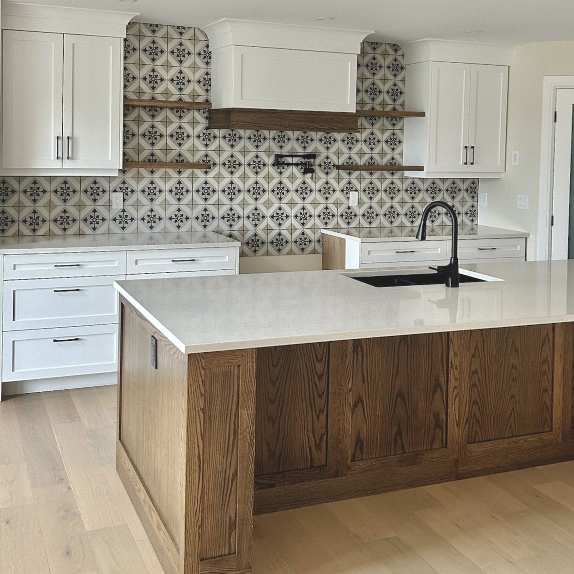 Elegant kitchen featuring white shaker cabinets, a patterned tile backsplash with a geometric design, and a wooden range hood.The island showcases a natural wood finish and a white countertop.