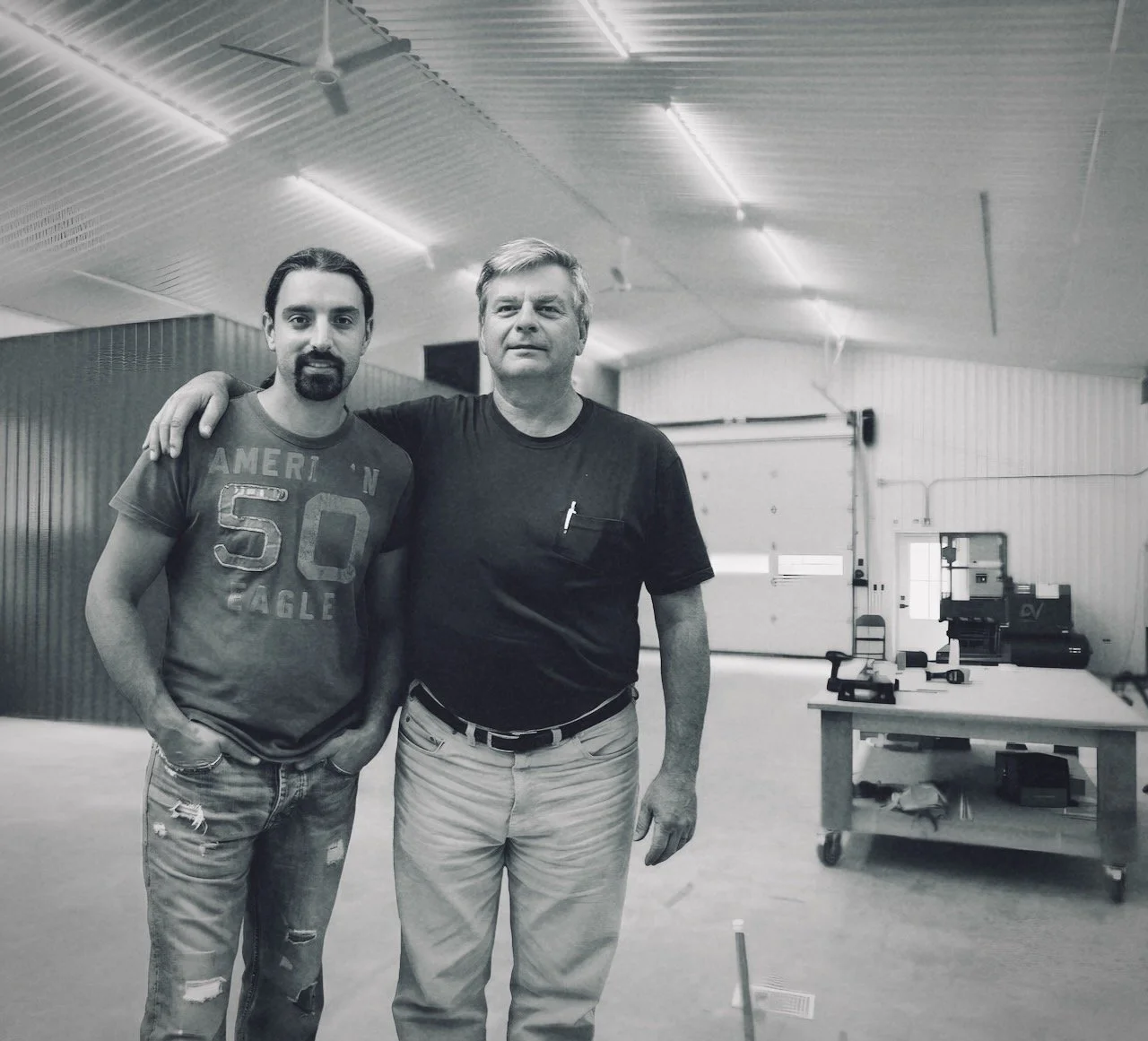 Two men standing inside a large industrial or workshop space, one with his arm around the other's shoulder, both looking at the camera.