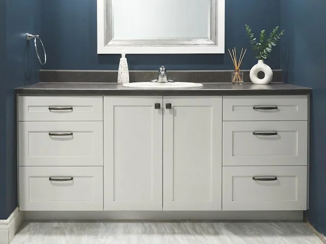 Bathroom vanity with white cabinets, a dark countertop, a mirror above, and decorative items including a soap dispenser, a plant, and reed diffuser.