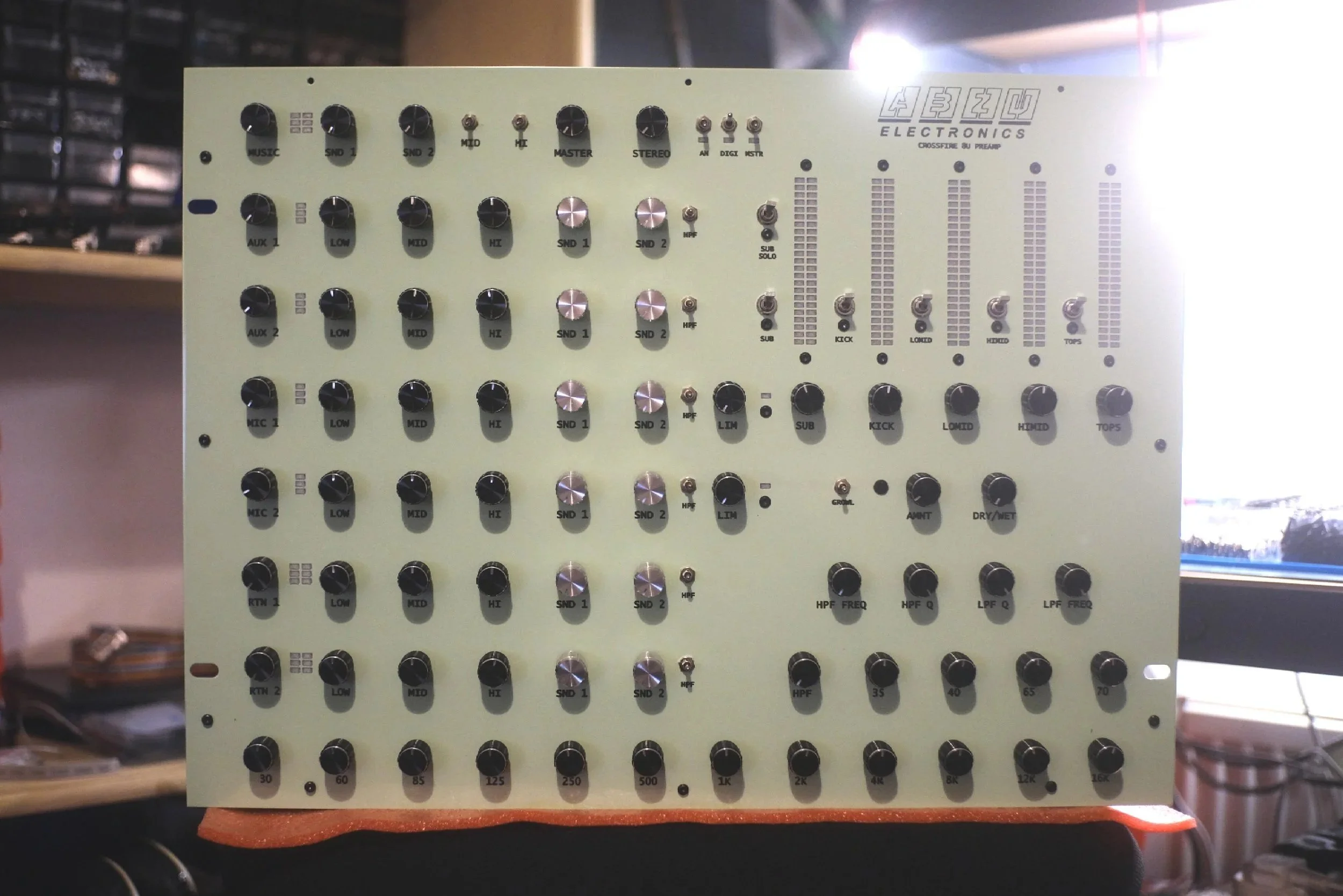 A beige electronic audio mixer with multiple knobs and sliders, placed on a surface in a room with shelves and equipment in the background.
