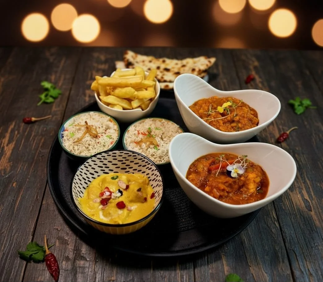 Thali platter with Indian dishes including two bowls of curry, a bowl of rice, a bowl of lentils, fries, and naan bread, all arranged on a black tray on a dark wooden table with a blurred string of lights in the background.