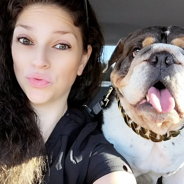 Woman with long dark curly hair and a bulldog with a gold chain collar sitting in a vehicle.