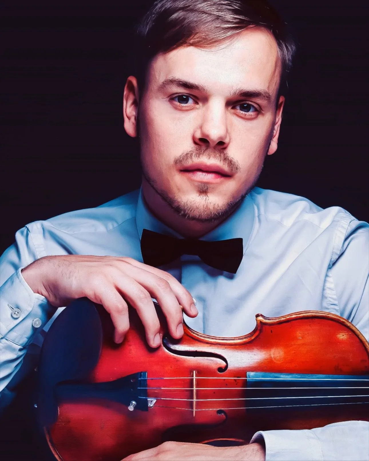 A young man with light skin, blue eyes, and light brown hair, wearing a white dress shirt and black bow tie, poses with a violin against a dark background.