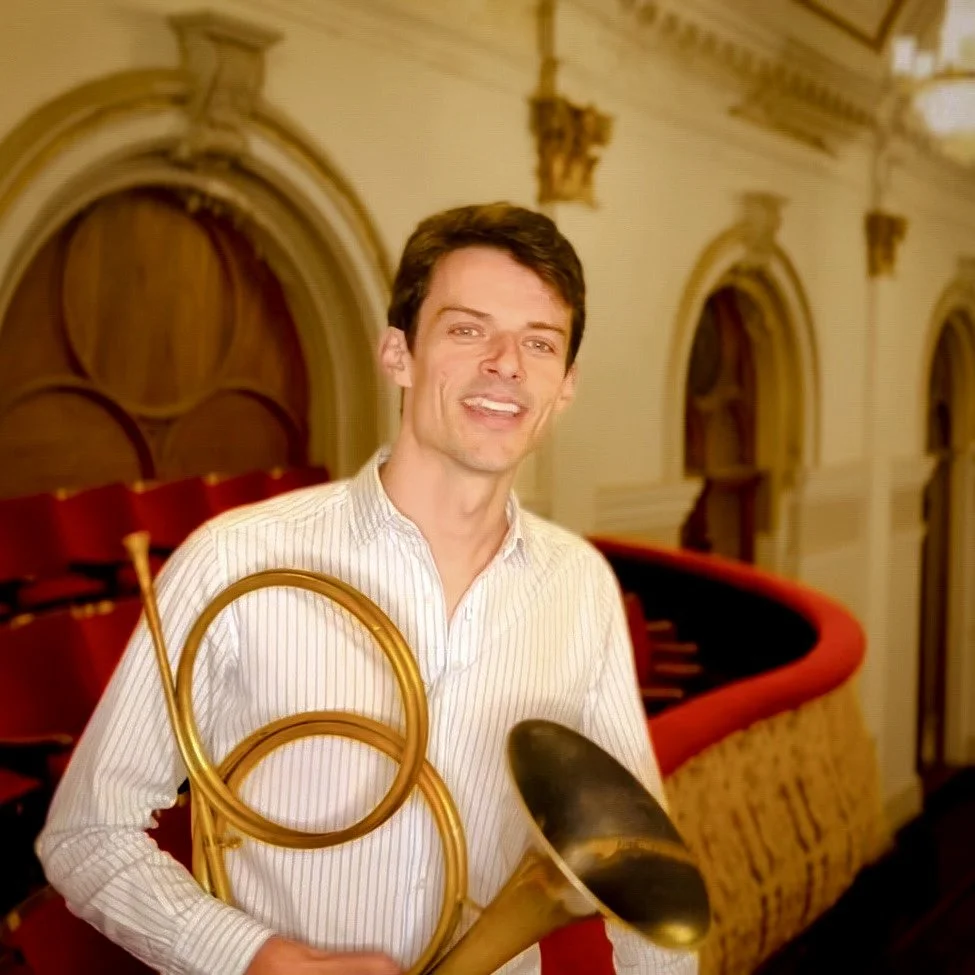 A man holding a horn in a theater with ornate architecture and red velvet seats.