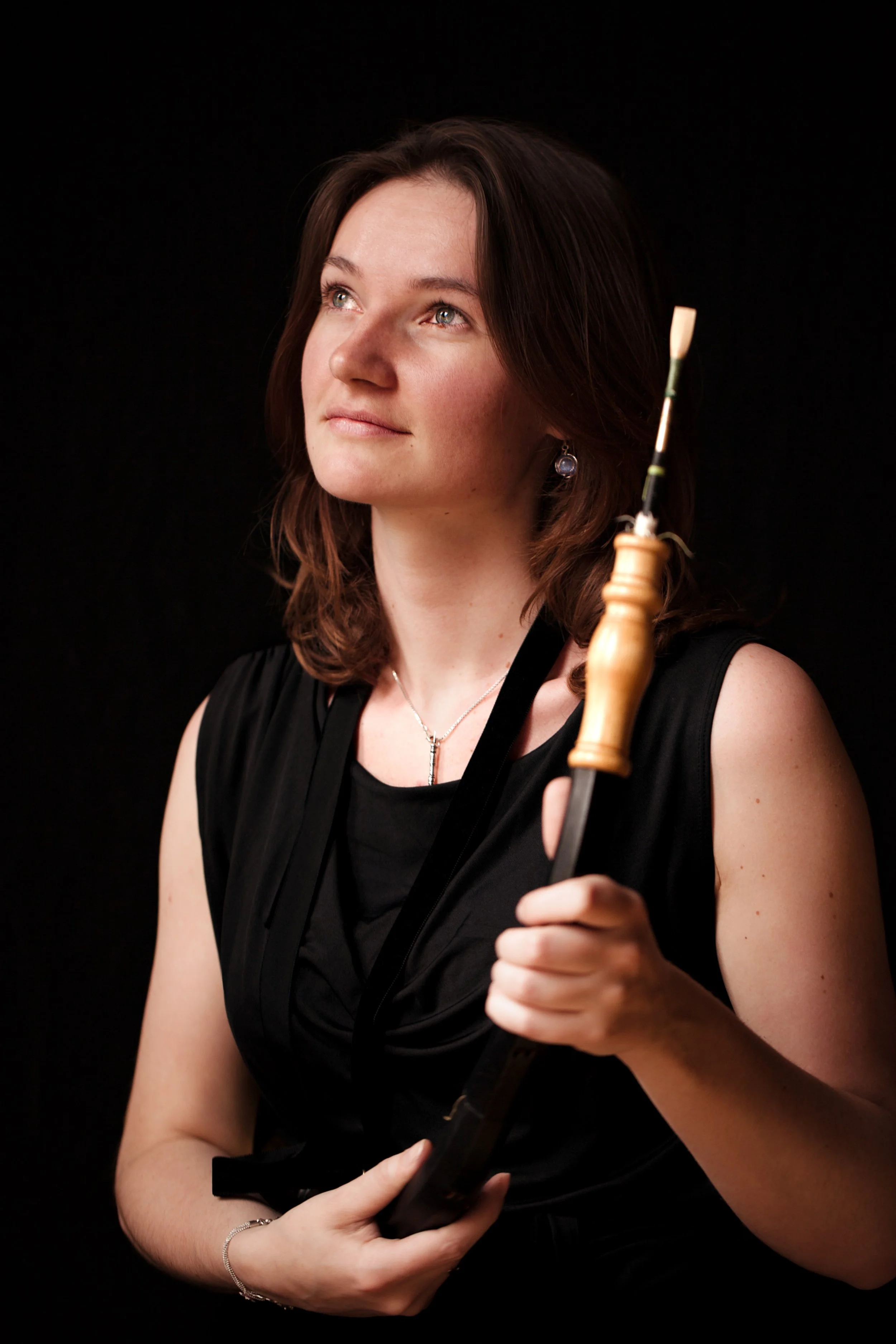 Young woman holding an oboe, wearing a black outfit, with a contemplative expression against a black background.
