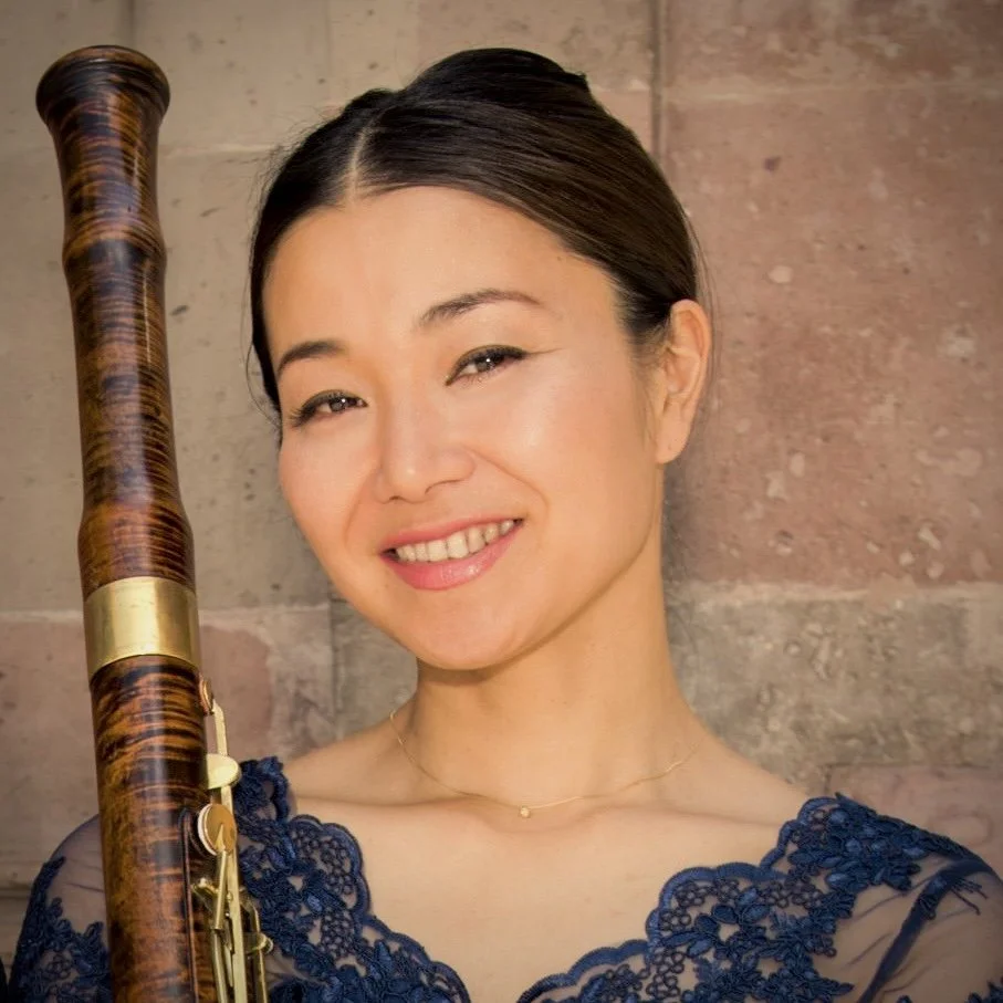A woman smiling, holding a bassoon, wearing a blue lace dress, with a brick wall background.
