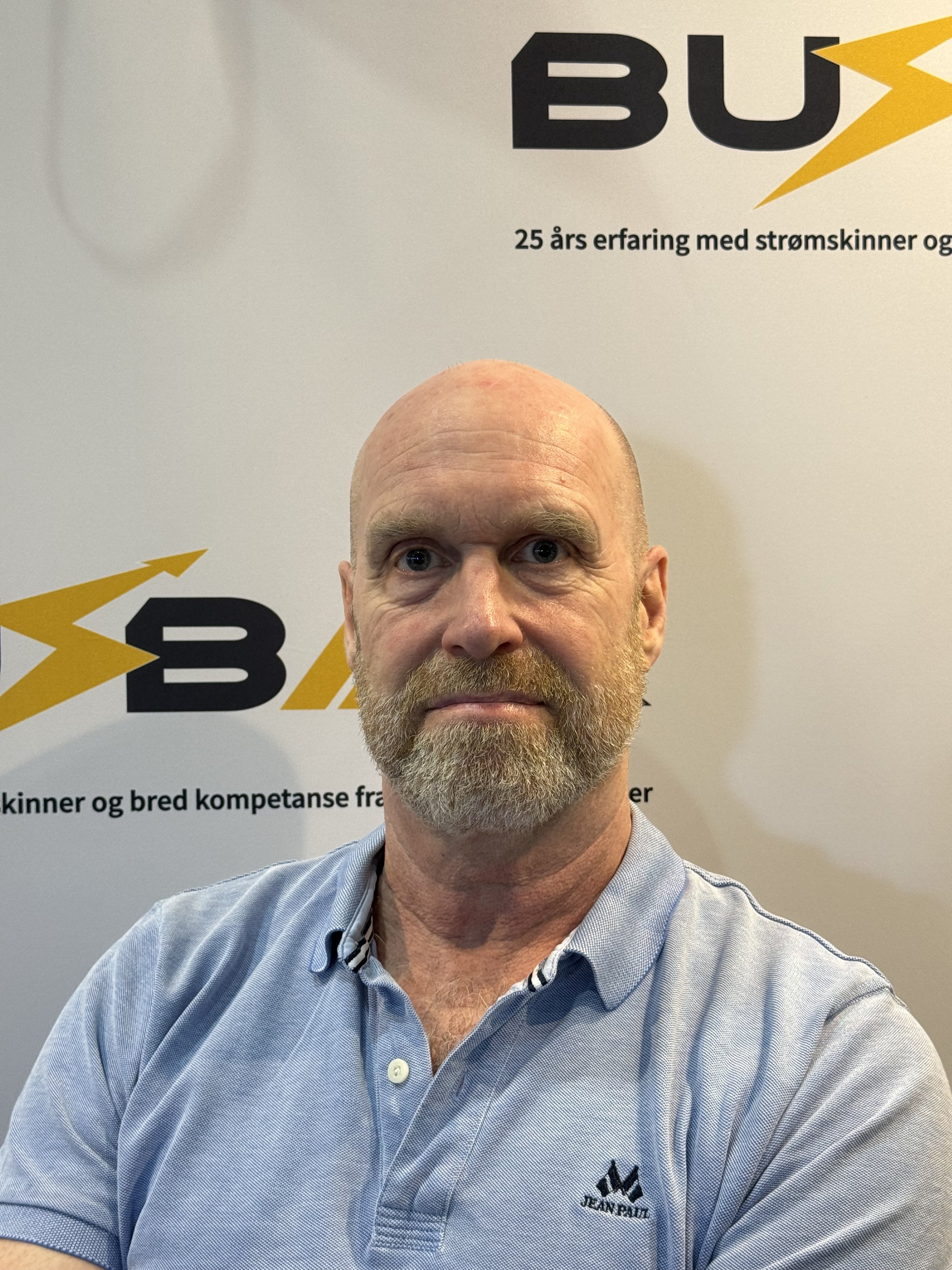 En mann med kort brun hår, skjegg og lyse klær står foran en vegg med en plakat som har norsk tekst og logo.