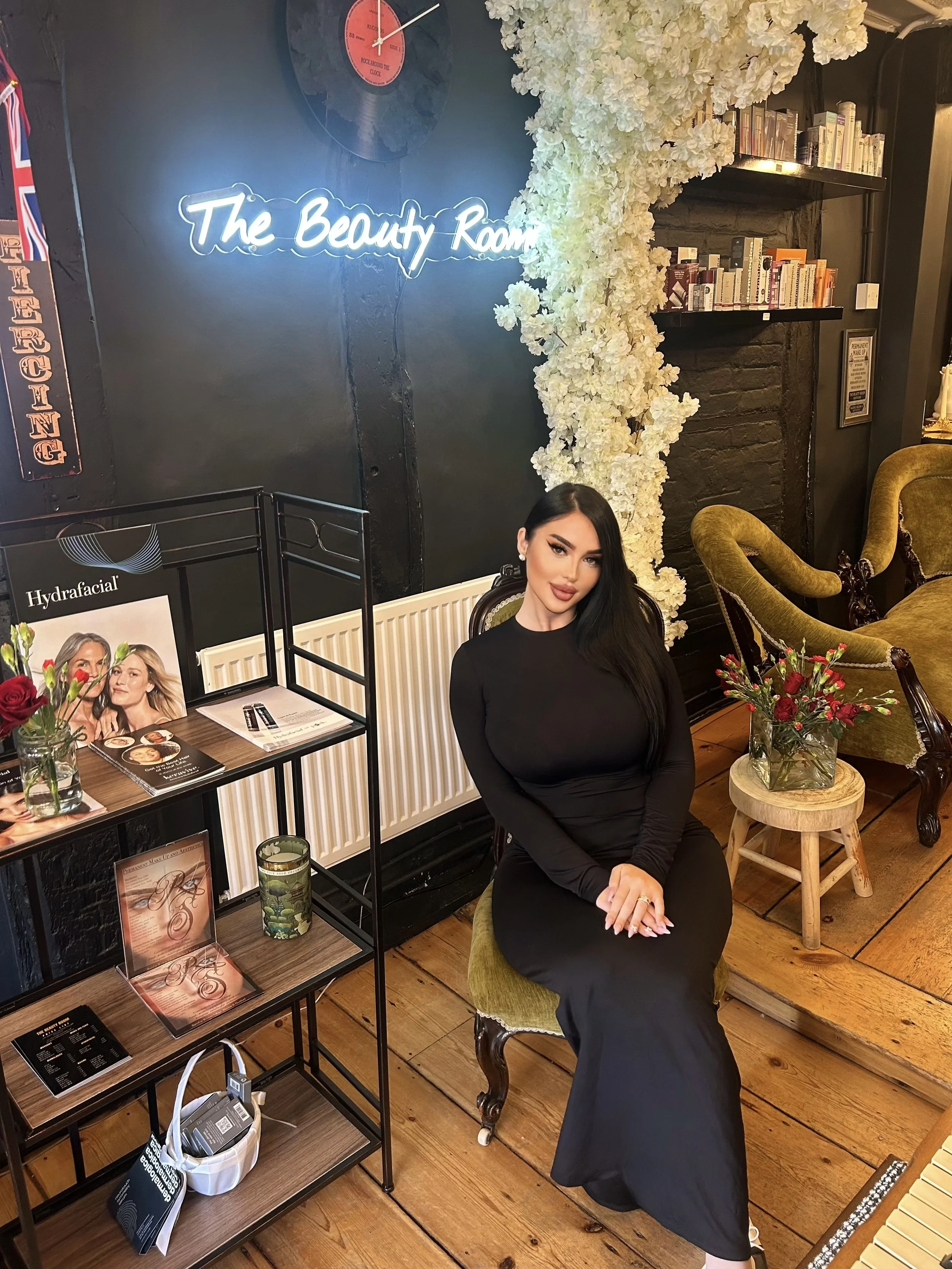A woman with long dark hair and wearing a black dress sits on a vintage-style green velvet chair in a room decorated with flowers, books, and a neon sign that reads "The Beauty Room."