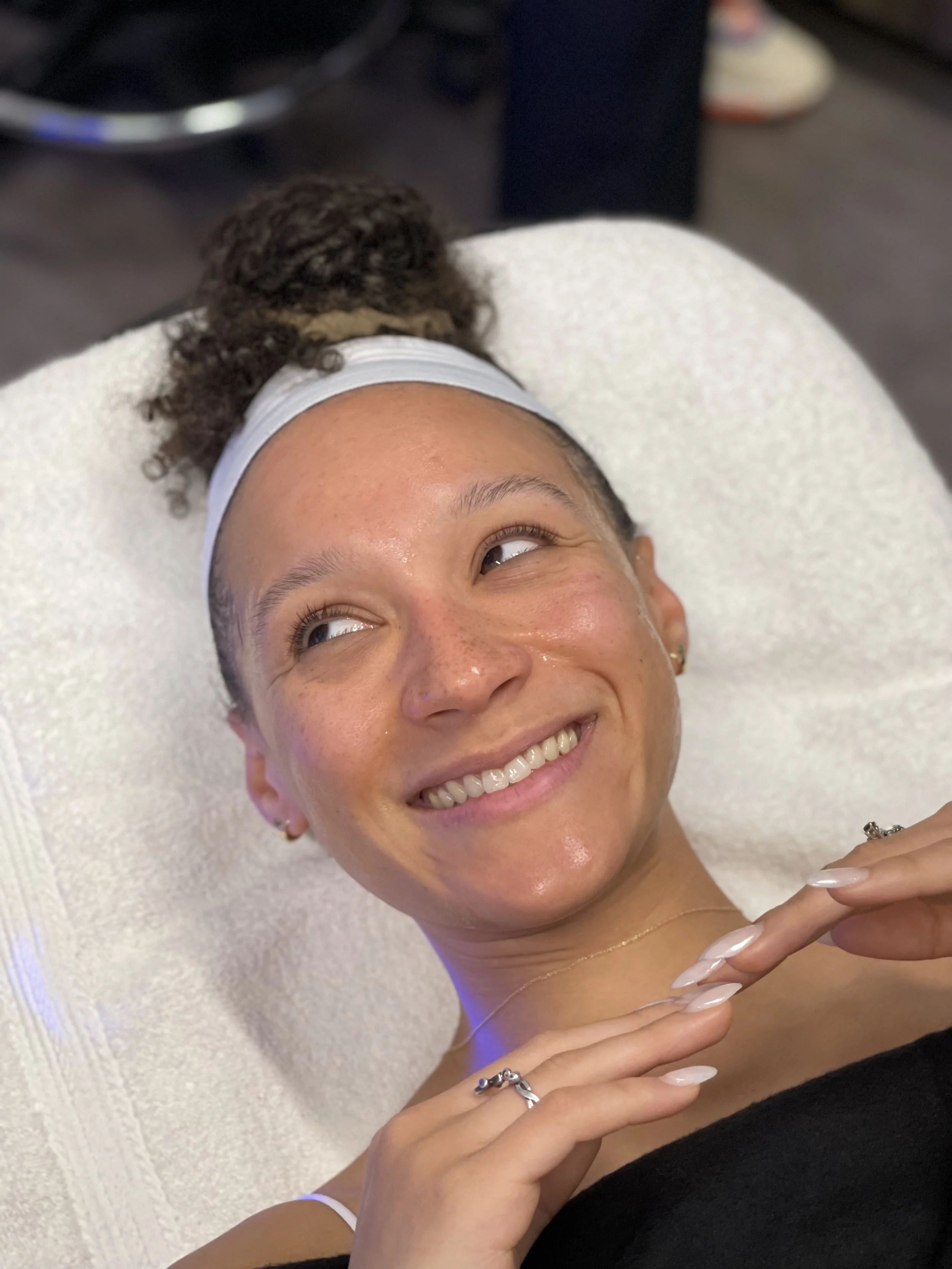 A woman with curly hair tied in a bun, lying on a white towel, smiling, with a shiny face. She is wearing earrings, a ring, a thin necklace, and a white headband.