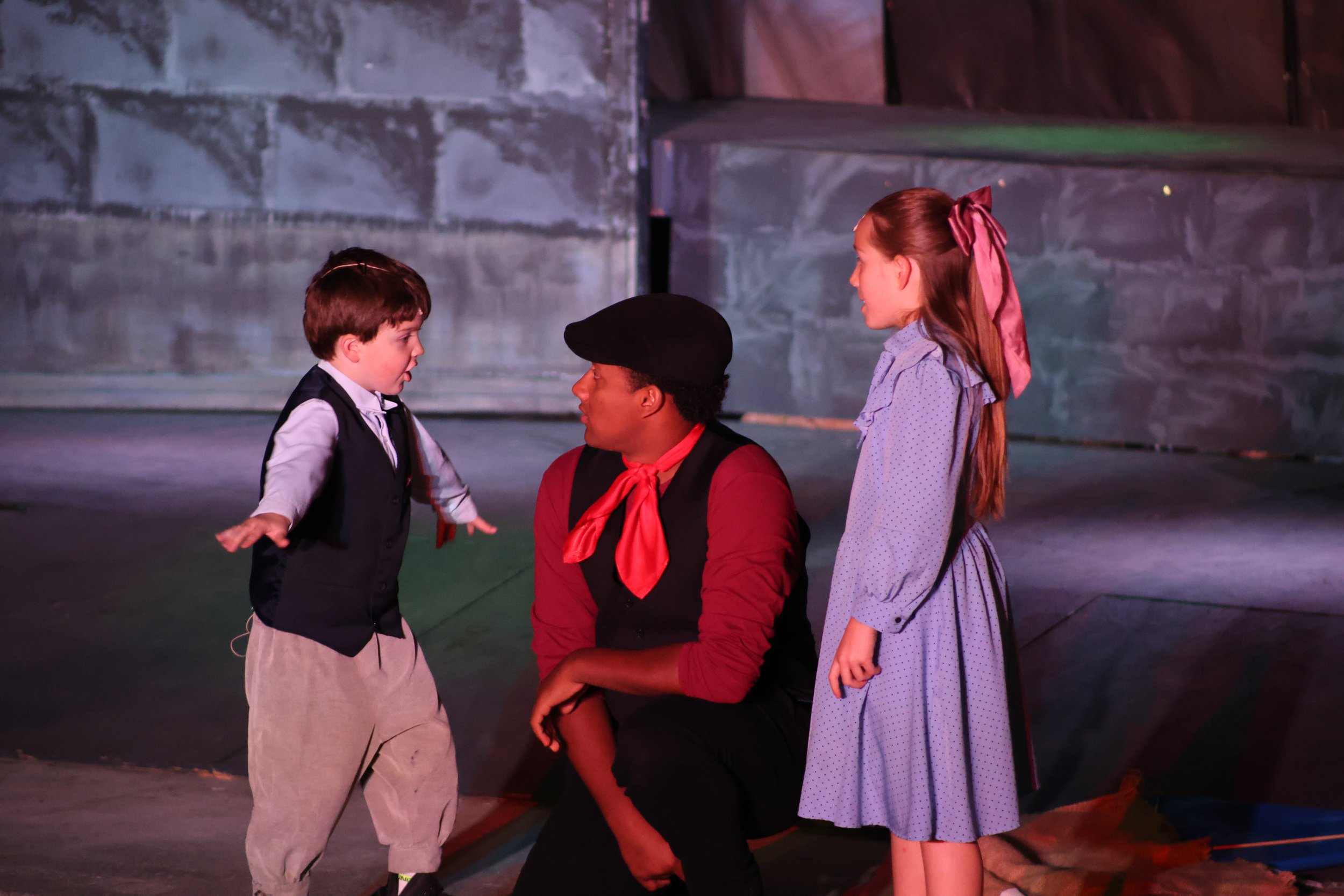 A young boy dressed in vintage clothing with a vest and khakis standing face to face with a man wearing a red scarf and a black beret, with two girls watching them, one girl has long hair tied with a pink ribbon and is wearing a purple dress. They ar
