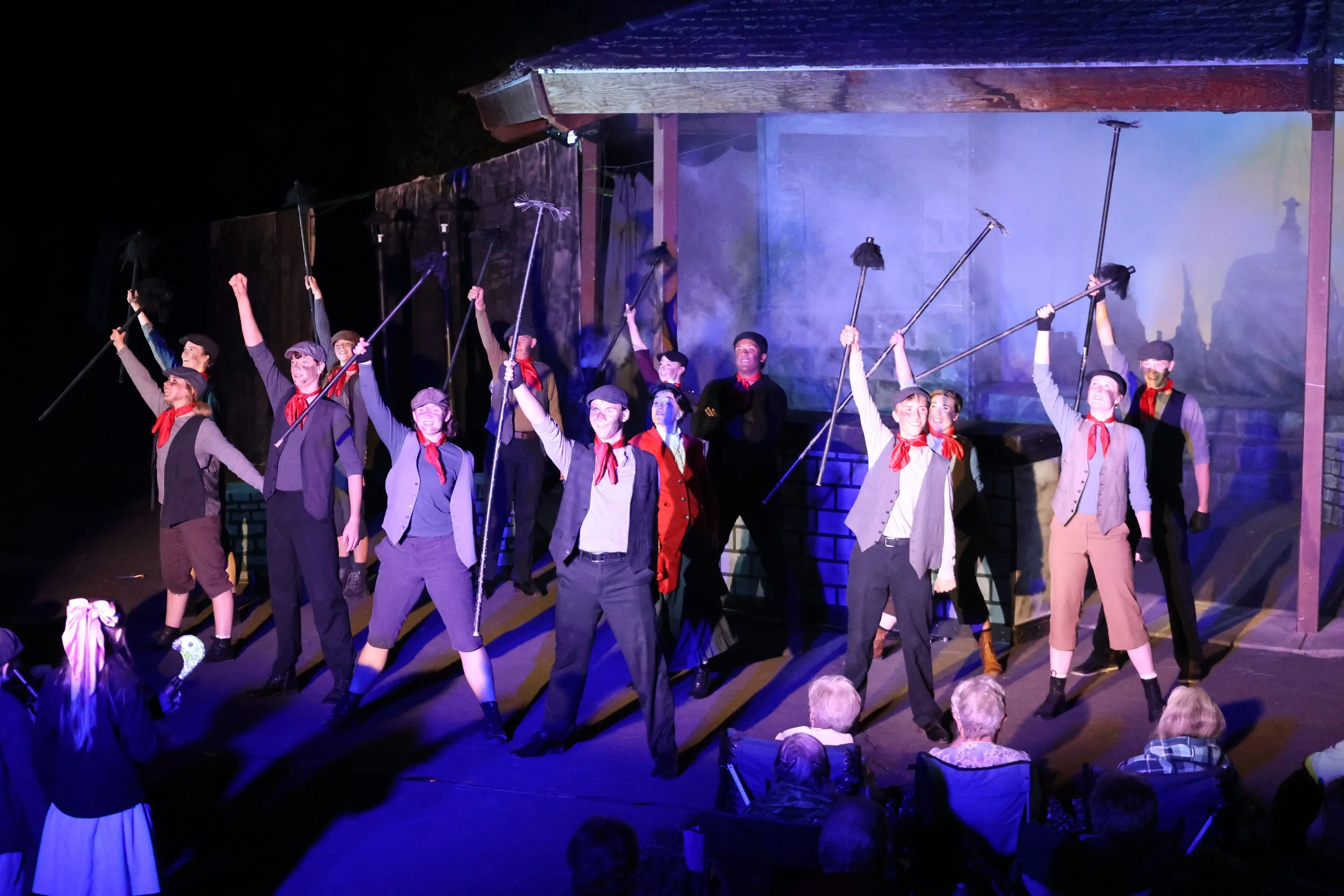 The image shows a theatrical stage performance with a group of actors dressed in historical costumes, raising their arms with sticks, likely portraying a scene with a sense of victory or celebration. The background includes a structure resembling a h