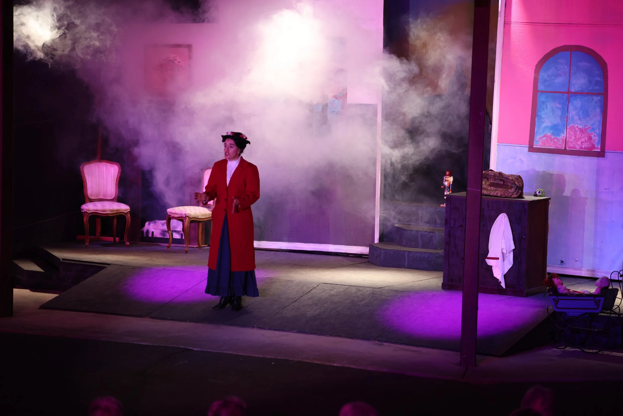 Woman in vintage costume performing on stage with pink chairs, a painted backdrop, and theatrical set pieces like a window and props, with stage fog.