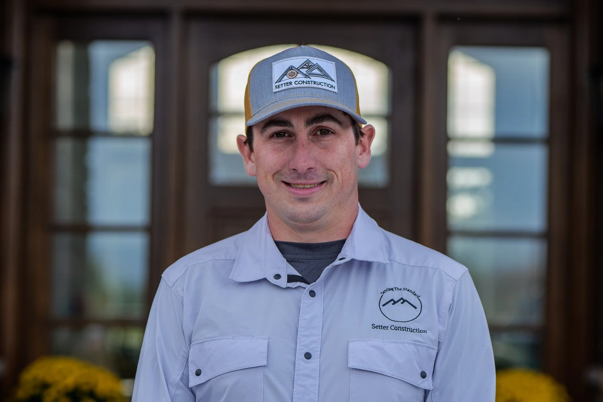 Portrait of a man wearing a construction company's uniform, including a gray cap with a logo and the words 'Setter Construction' and a light-colored shirt with the company's logo, standing indoors with windows and yellow flowers in the background.