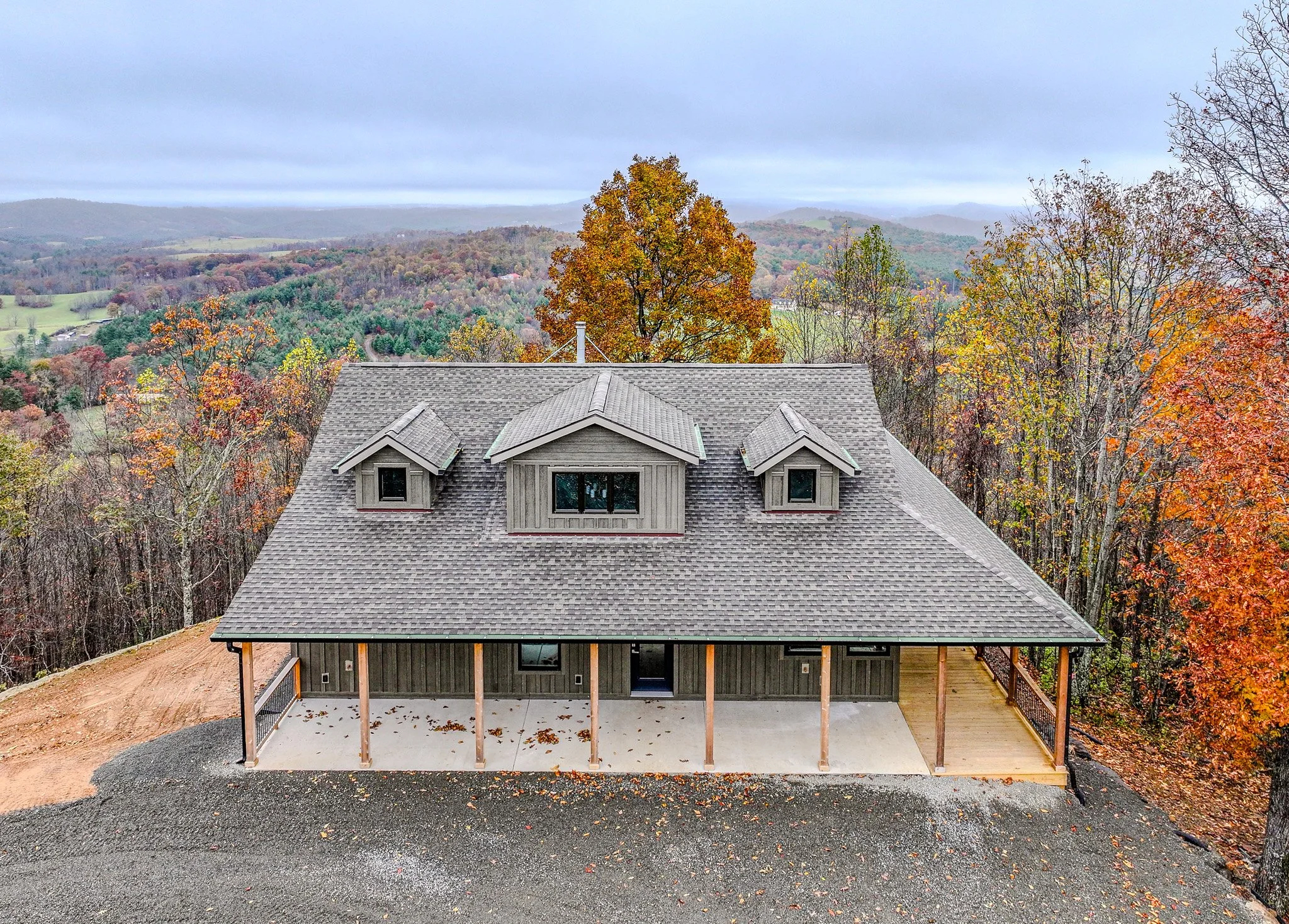 A two-story house with a gray exterior, large front porch, and dormer windows on the roof, surrounded by autumn trees with orange and yellow leaves, under a cloudy sky.