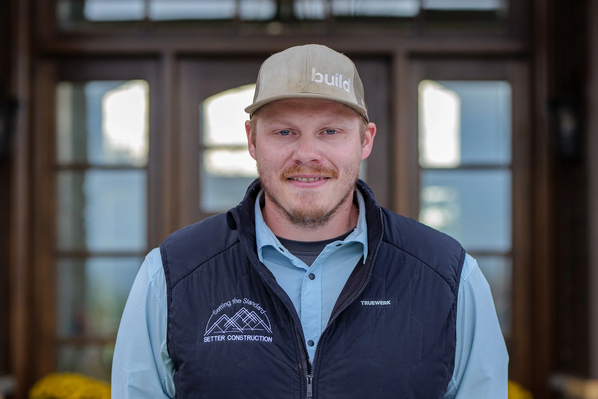 A man wearing a beige baseball cap with 'build' embroidered on it, a light blue button-up shirt, and a navy vest with the logos 'Setter Construction' and 'TRUEWERK', standing indoors in front of a wooden window frame.