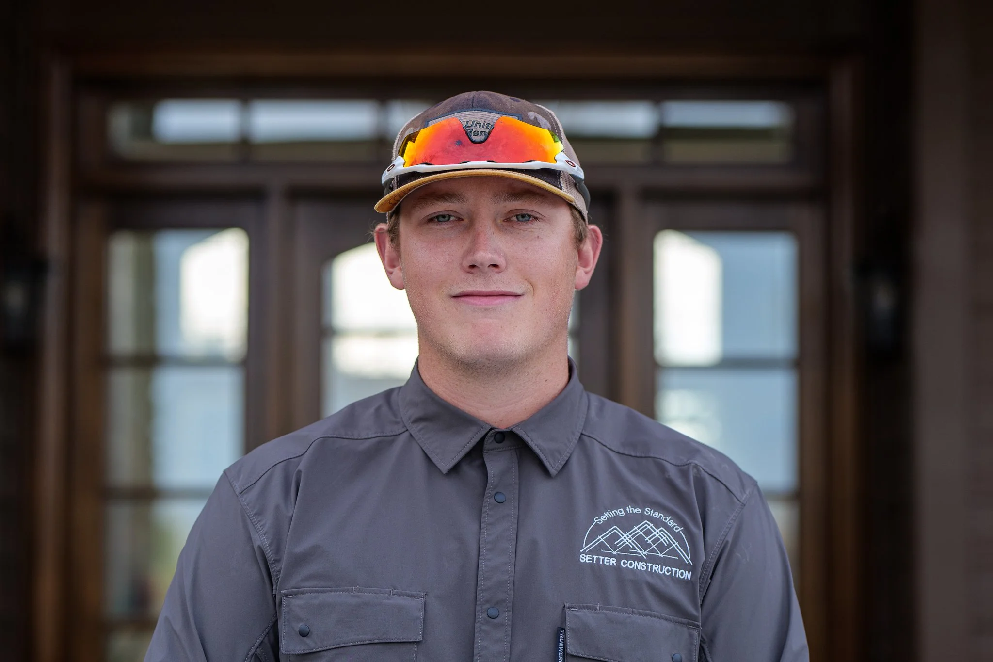 A young man wearing a gray shirt with a logo, a cap, and sunglasses on his head, standing indoors in front of a windowed background.