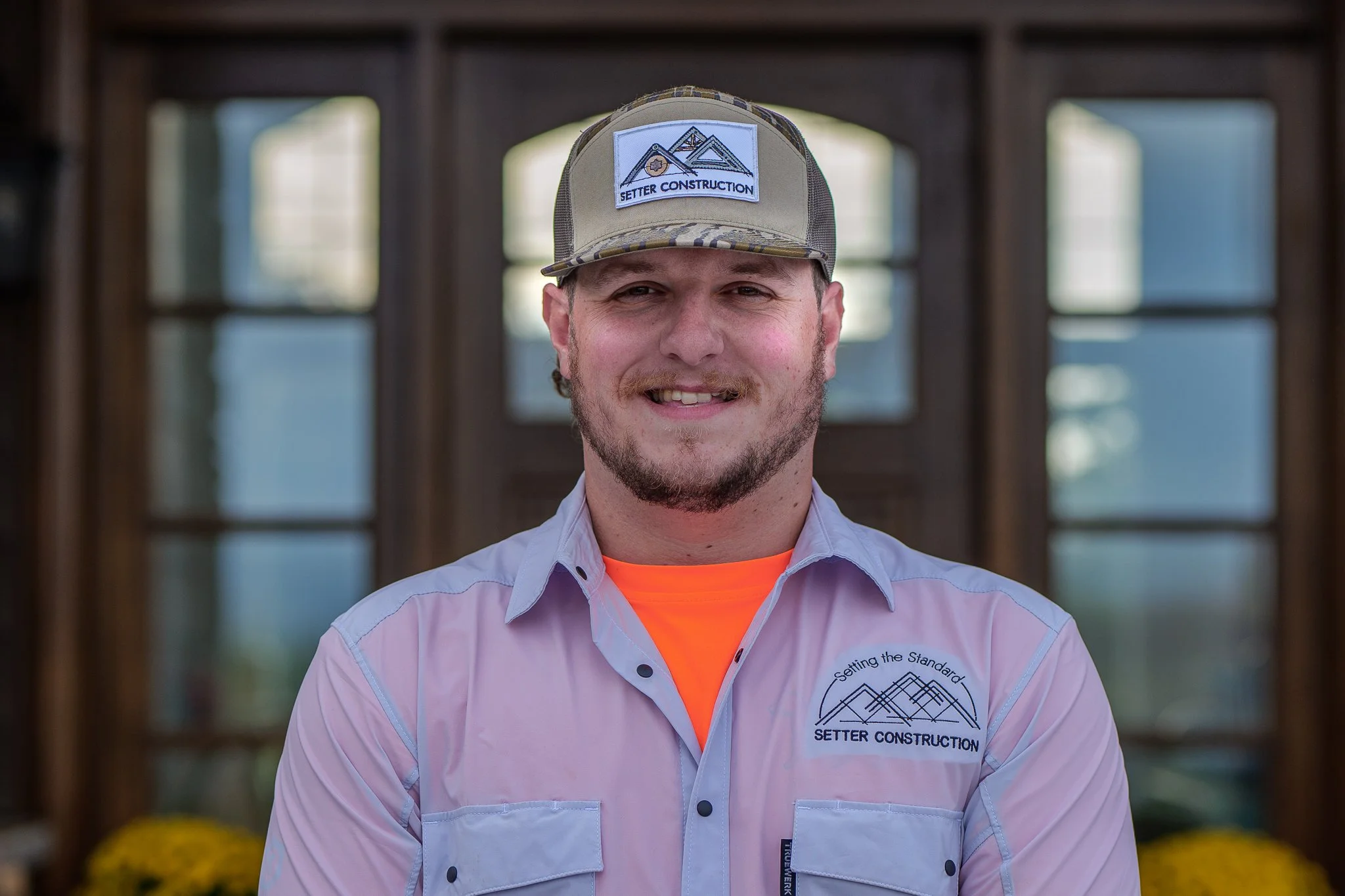 A man wearing a plaid cap with a logo that says 'Setter Construction', a light pink uniform shirt with the same logo, and an orange shirt underneath, standing in front of a wooden structure with large windows.