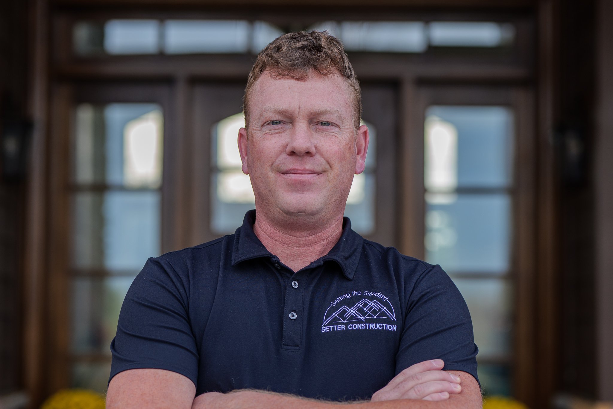 A man standing indoors in front of large windows with wooden frames, crossing his arms, wearing a navy blue polo shirt with a logo that reads 'Setter Construction' and a mountain design.