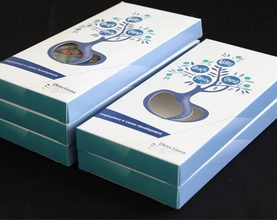 Three stacked boxes with a tree logo and a cutout window on the top, containing colorful game pieces inside, against a black background.