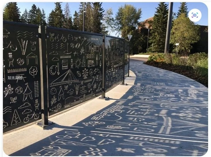 Outdoor pathway with decorative black metal fence and shadows cast on the sidewalk, surrounded by trees and greenery.