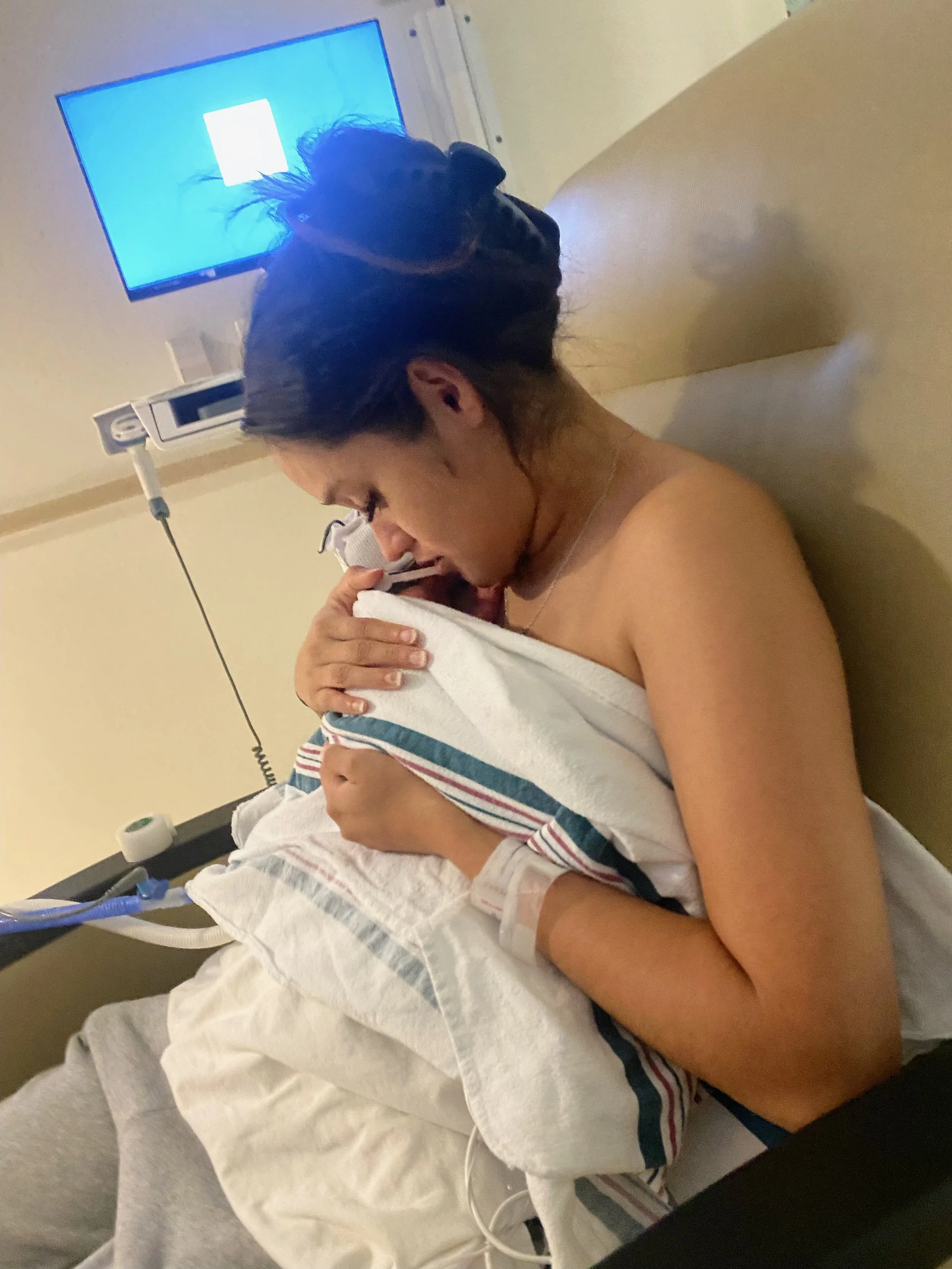A mother in a hospital chair cradles a newborn baby wrapped in a blanket, both resting peacefully.