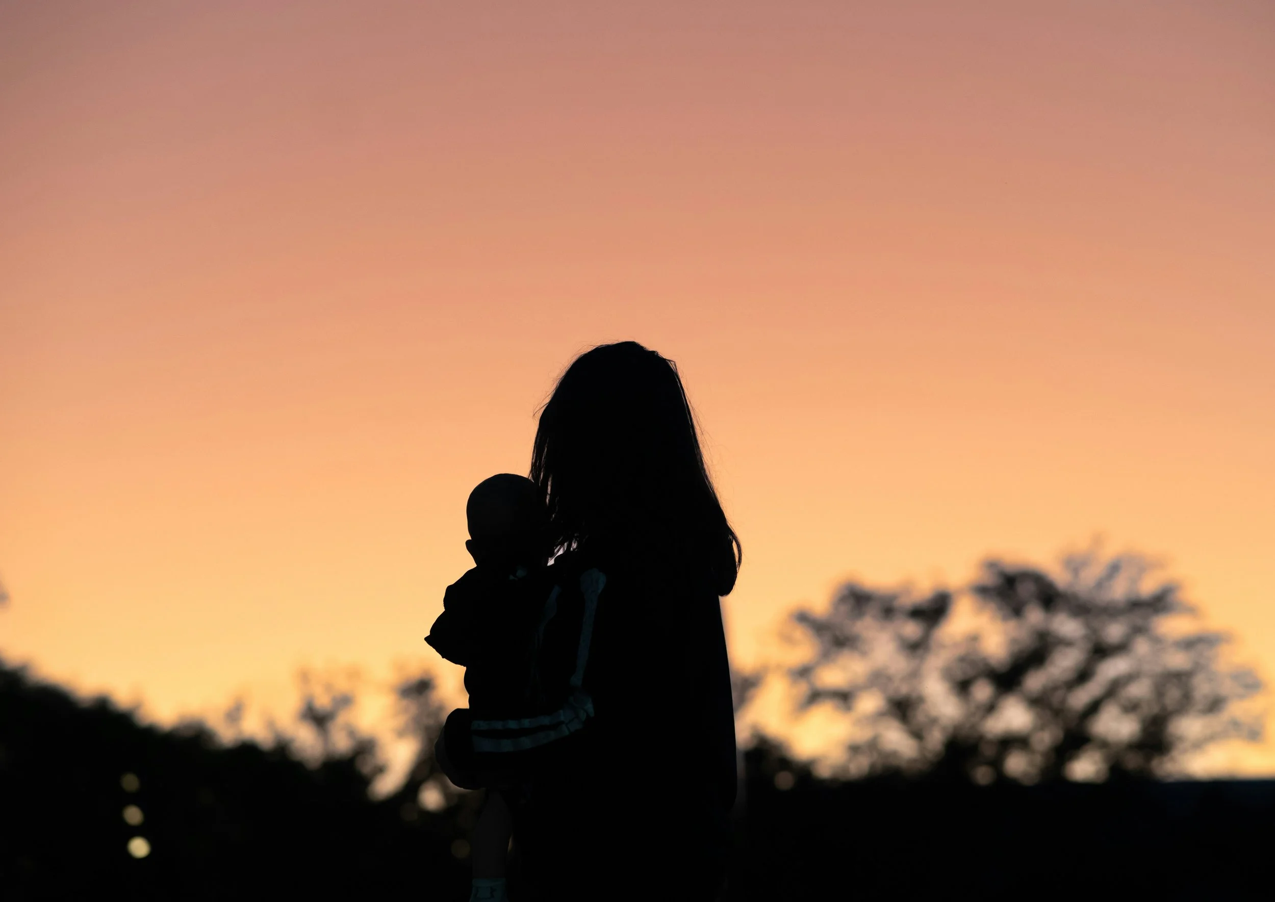 Silhouette of a mother holding a baby against an orange sunset sky.