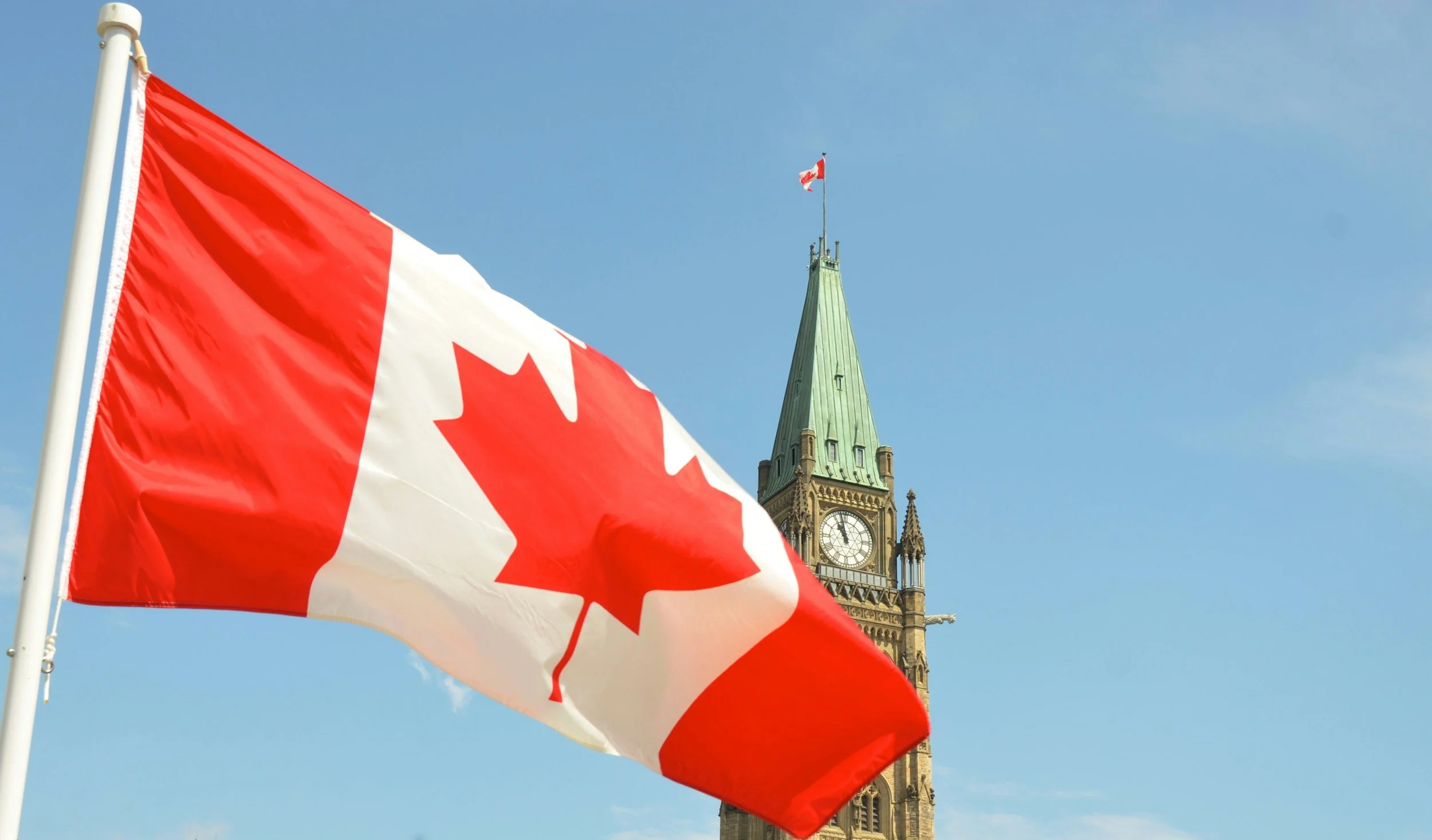 A Canadian flag proudly waves before the Peace Tower on Parliament Hill in Ottawa beneath a clear blue sky.