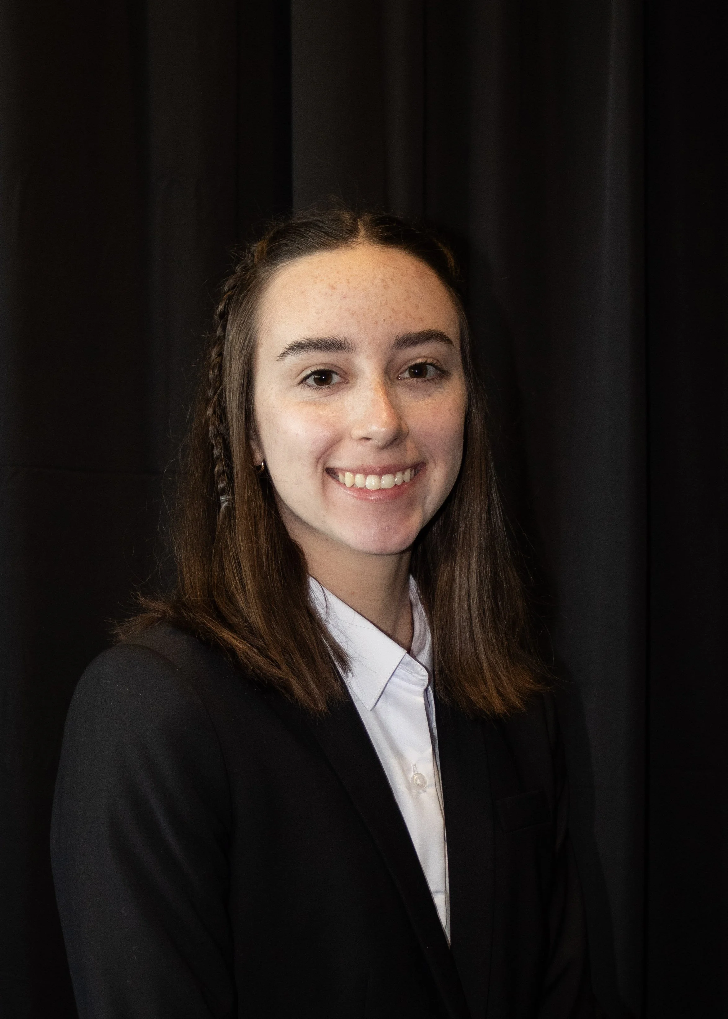 Emma Powell smiles for a professional headshot, wearing a black blazer and white button-down shirt against a dark background.