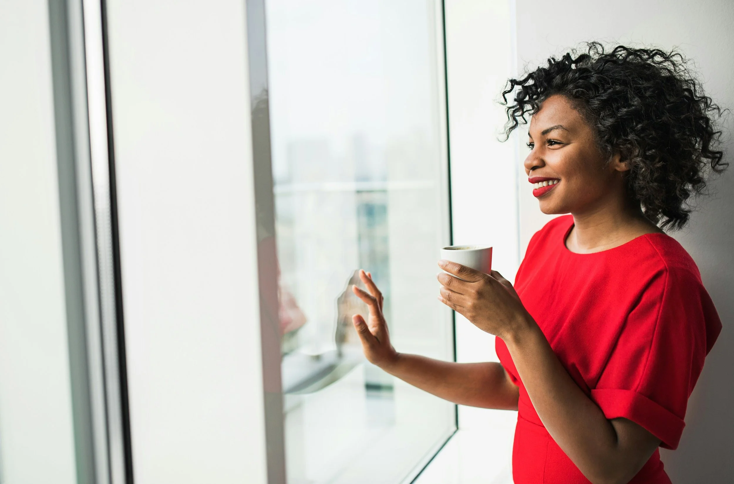 A woman stands quietly by a window holding a warm mug, taking a calm moment to herself.