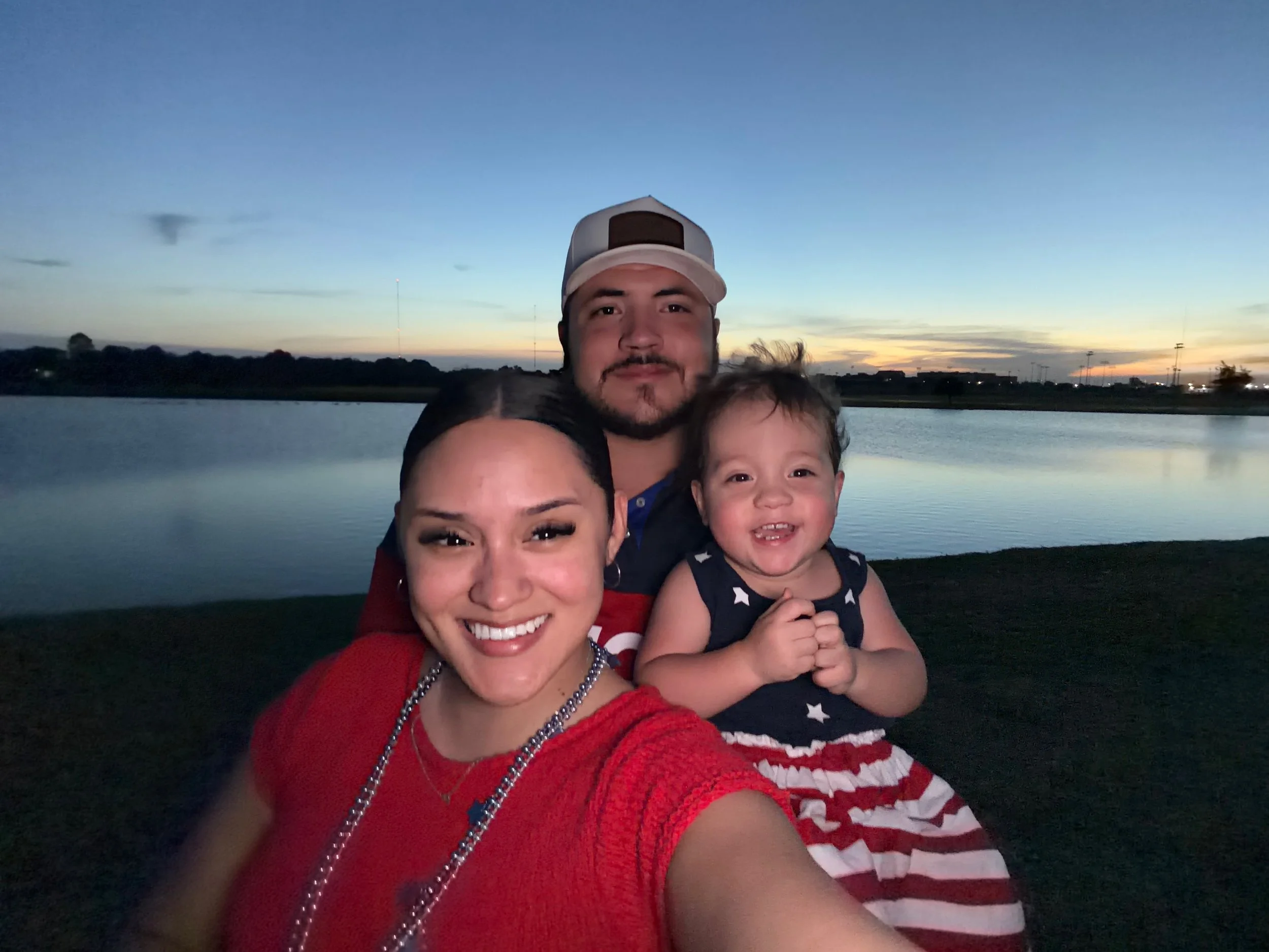 Family of three smiling by a lake at sunset; child wears a star-spangled dress, mother in red, father in a cap.