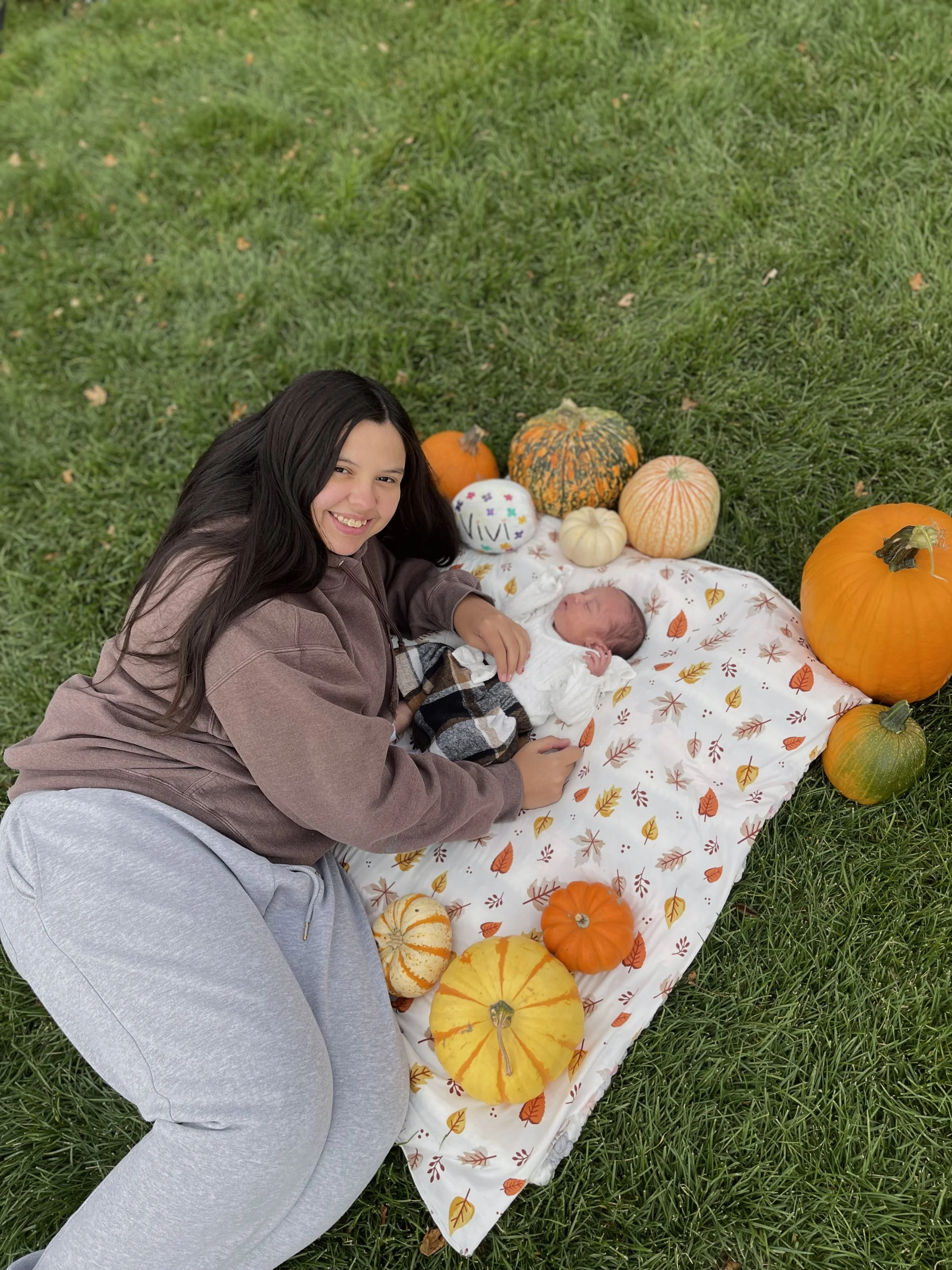 A smiling young mother lies on a blanket surrounded by pumpkins, holding her newborn baby outdoors on green grass.