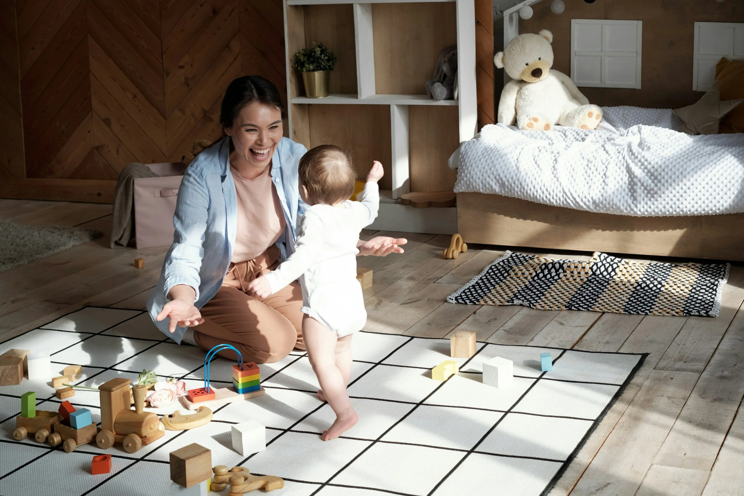 A mother encourages her baby as they take steps toward her on a playroom floor.